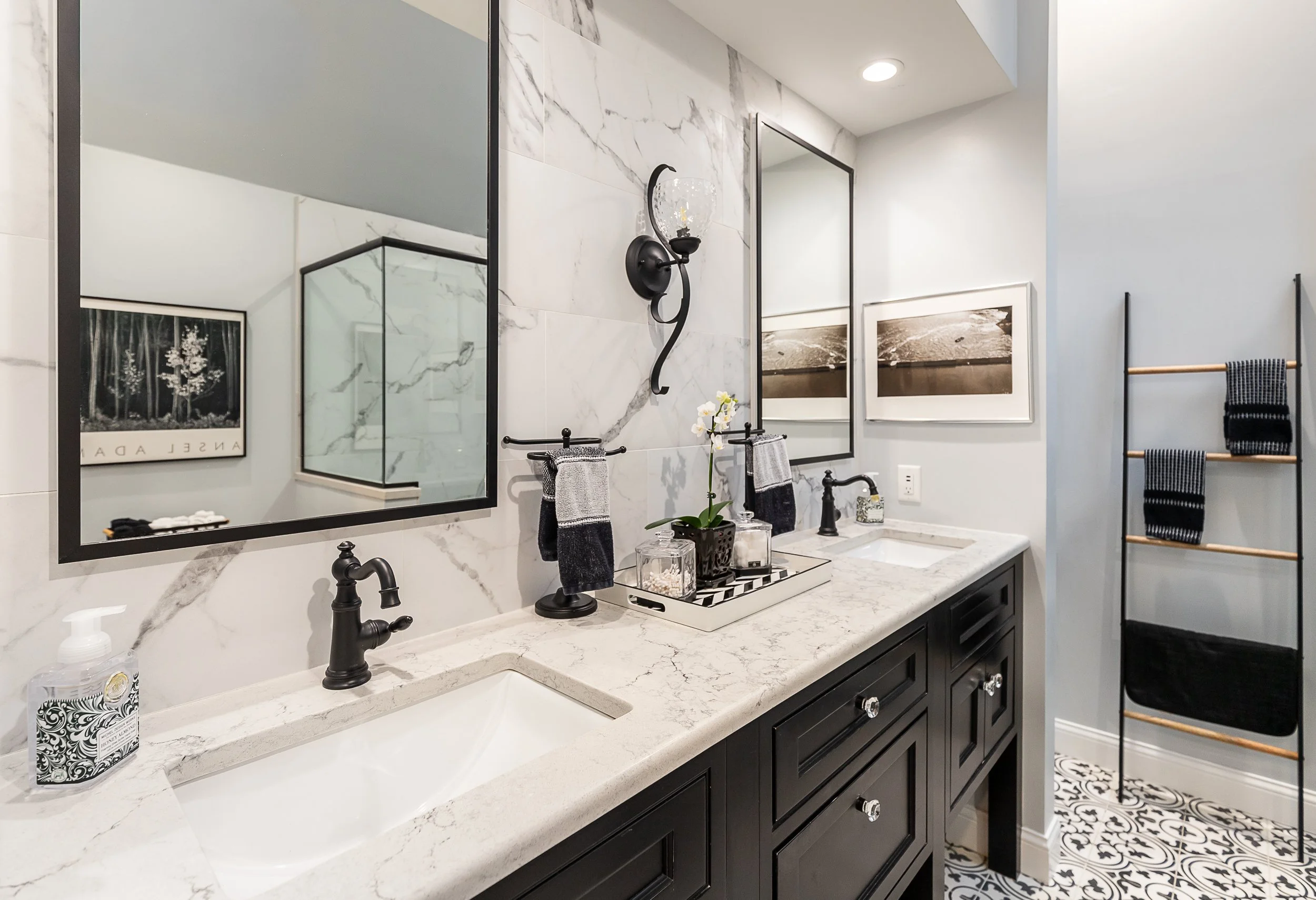 Modern bathroom with a double vanity, black fixtures, quartz countertops, framed mirrors, black and white artwork, hand towels, decorative accessories, and patterned tile floor.