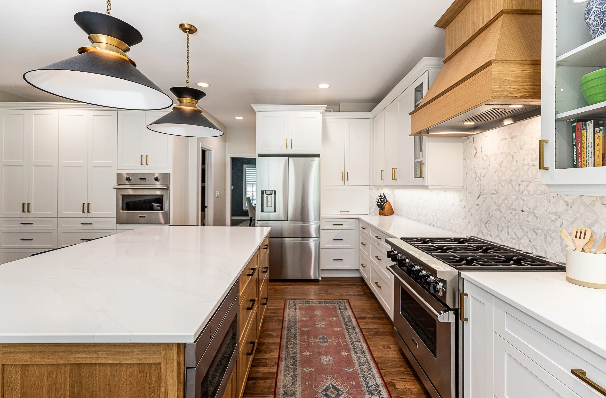 Modern kitchen with white cabinets, stainless steel appliances, a large island with a quartz countertop, a red patterned rug, and a wooden range hood.