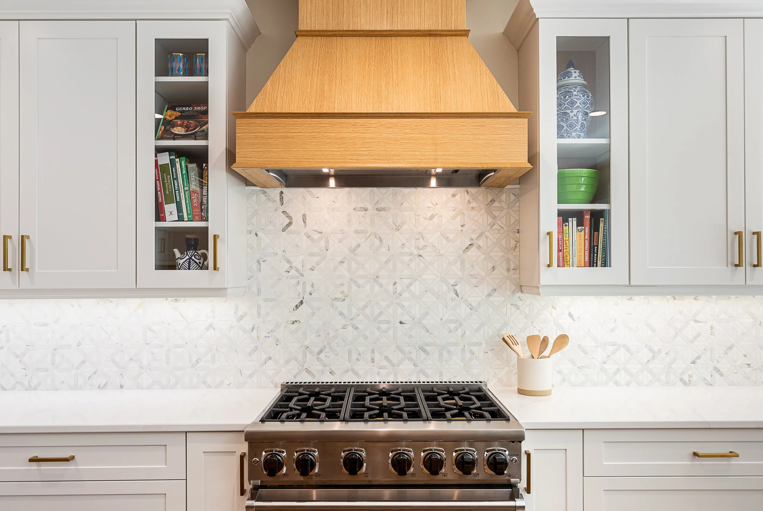 Modern kitchen with white cabinets, gold handles, a stainless steel stove, open shelving, decorative backsplash, and a wooden range hood.