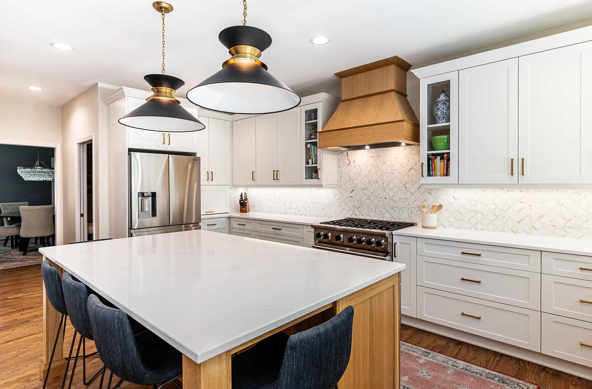 Modern kitchen with white cabinets, stainless steel refrigerator, wooden range hood, black and gold pendant lights, and a large white island with black chairs.
