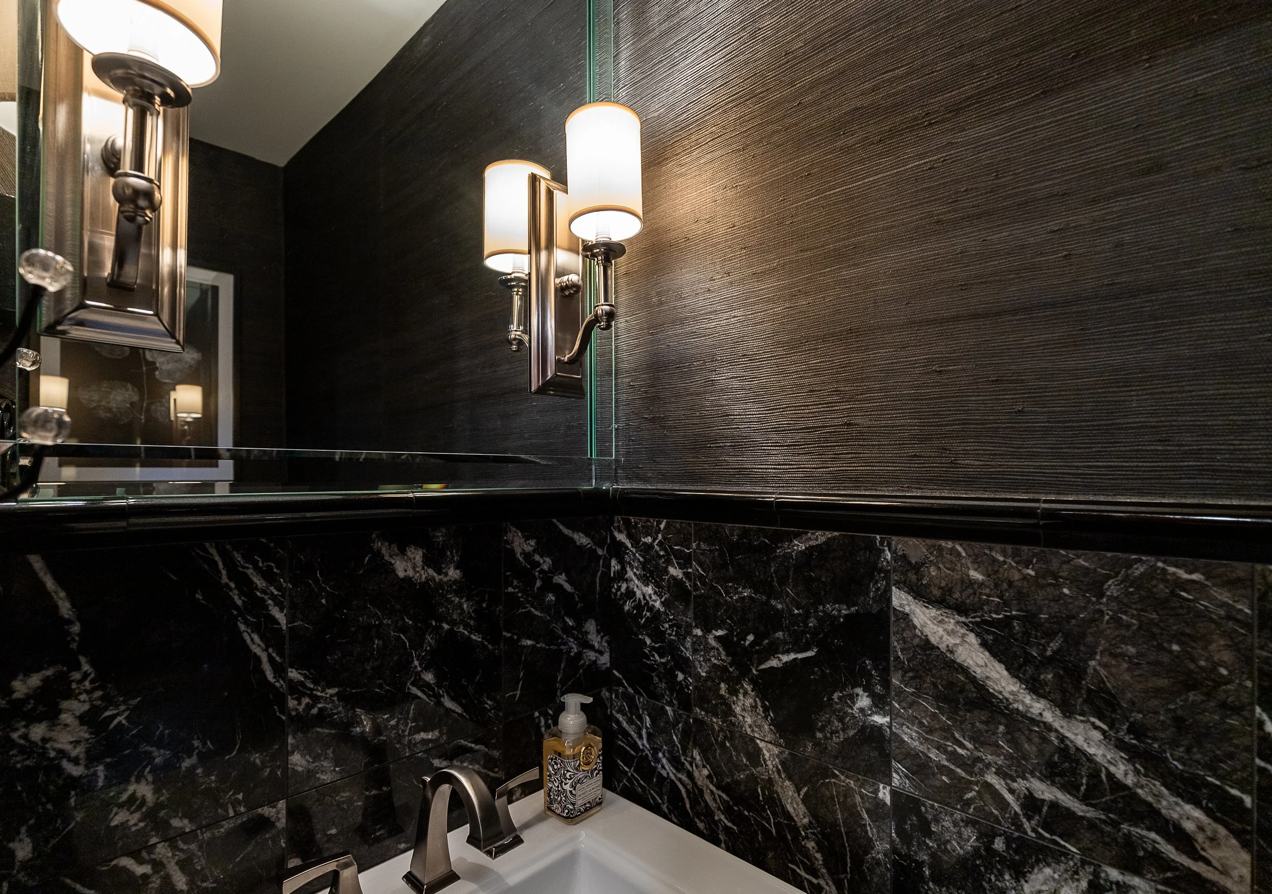 A bathroom with black marble tiles on the lower half of the wall, a white sink with a soap dispenser, and dark wood paneling on the upper wall. There is a mirror above the sink and two wall-mounted light fixtures with white shades.
