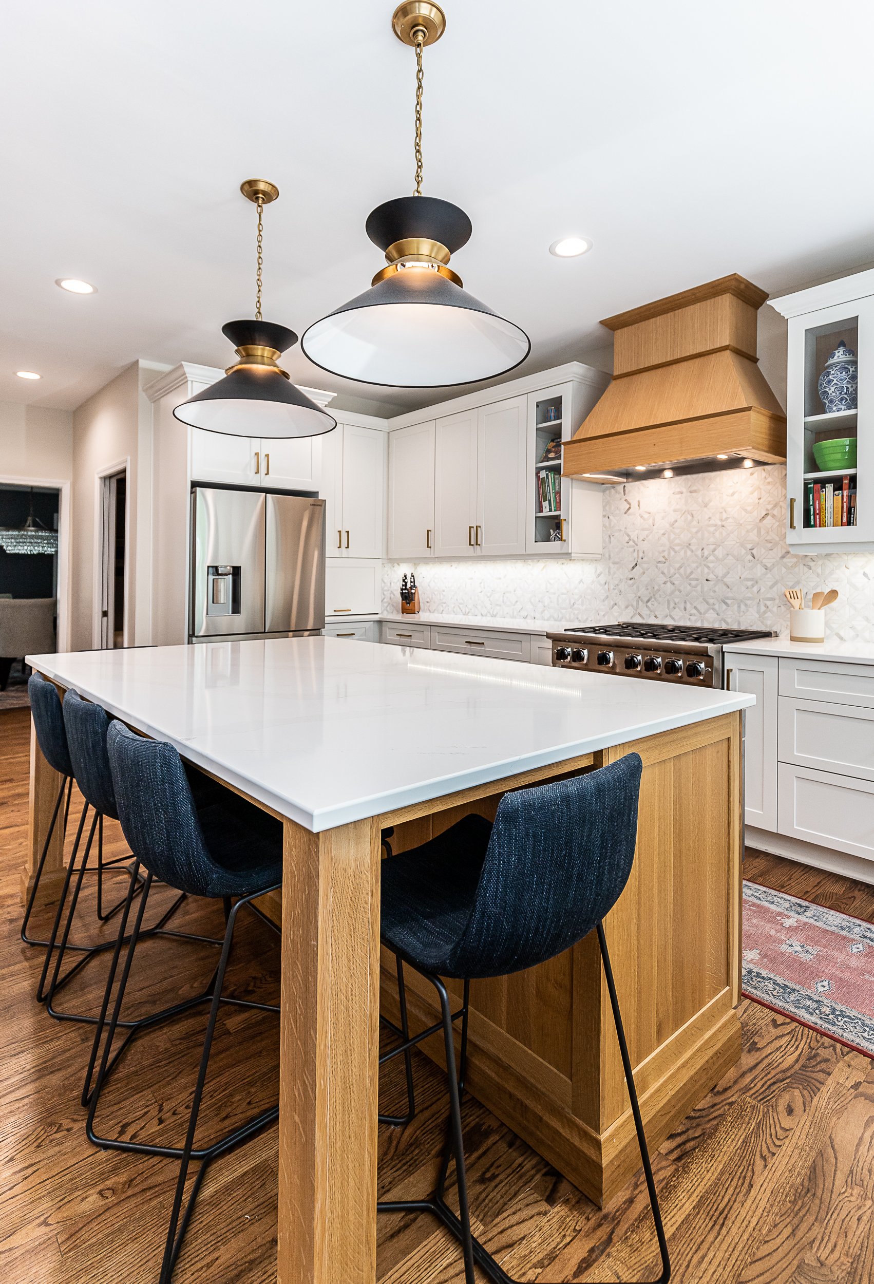 Modern kitchen with white cabinets, a large wooden island with a white countertop, black bar stools, stainless steel refrigerator, a gold and black range hood, and black pendant lights hanging from the ceiling.