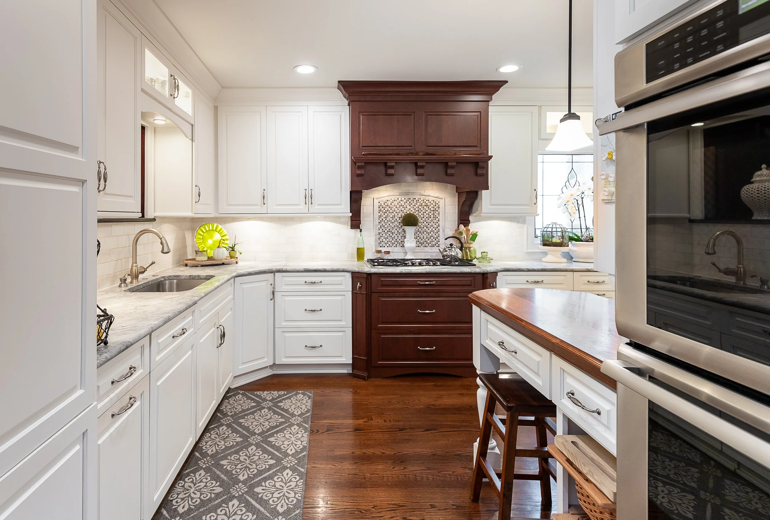 Kitchen with white cabinets, a brown range hood, quartz countertops, and hardwood floors. Small decorative items and plants are on the counters, and a patterned gray rug is on the floor.