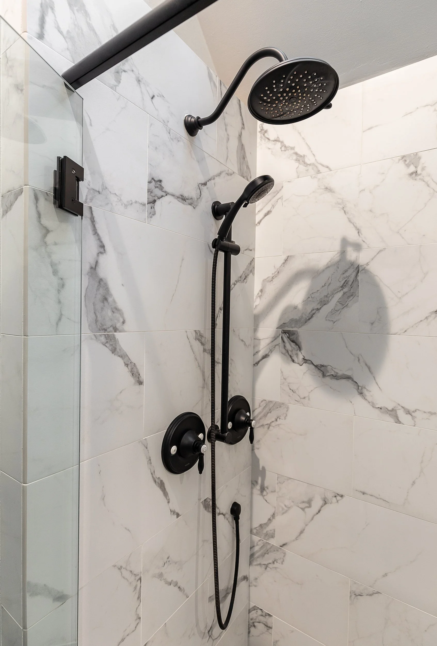 Black shower fixtures installed on a white marble-looking tile wall with gray veining, including a rainfall showerhead, a handheld showerhead, and control knobs.