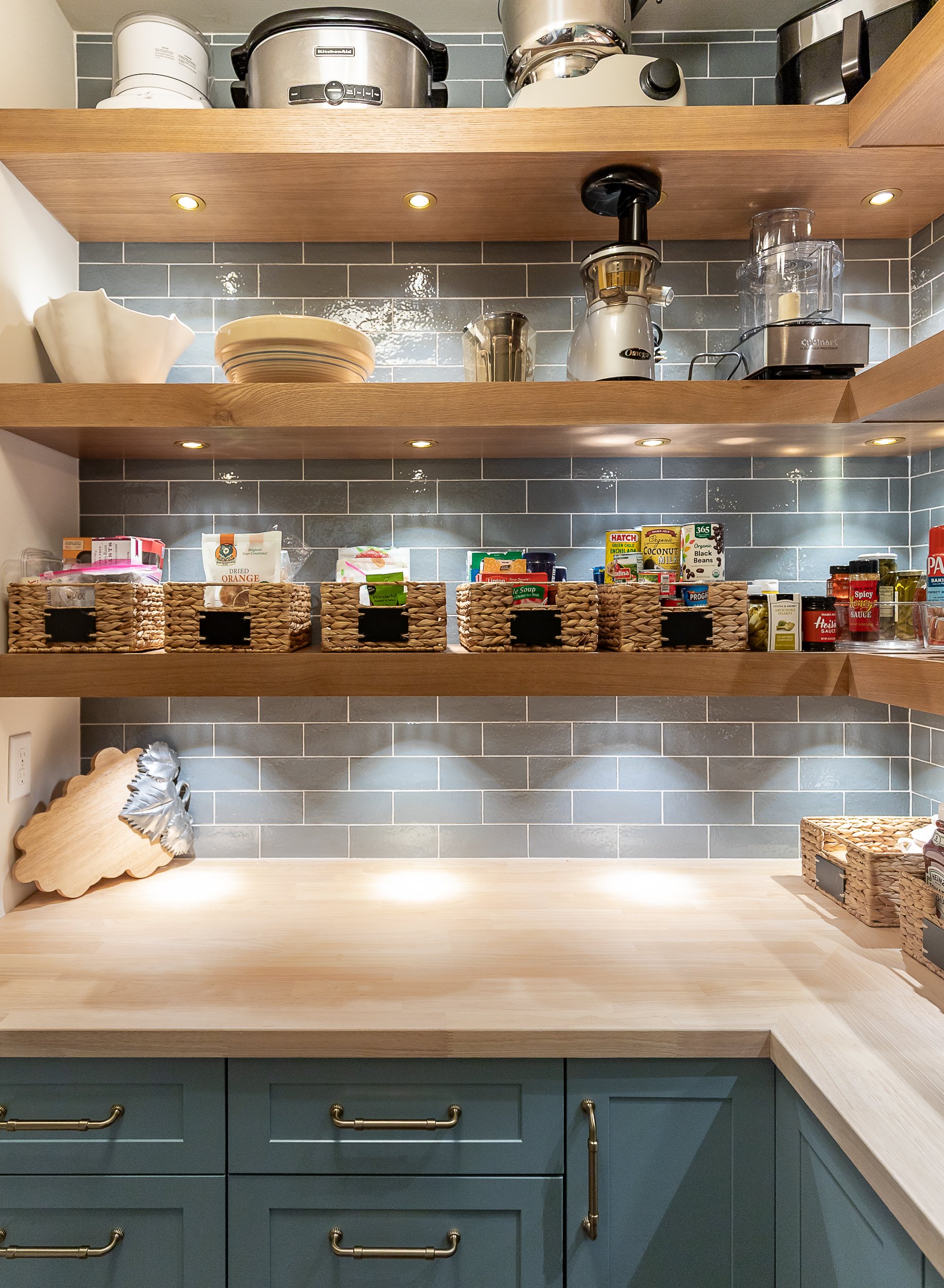 Open kitchen shelving with various appliances, dishes, food items, and baskets, with a gray tile backsplash.