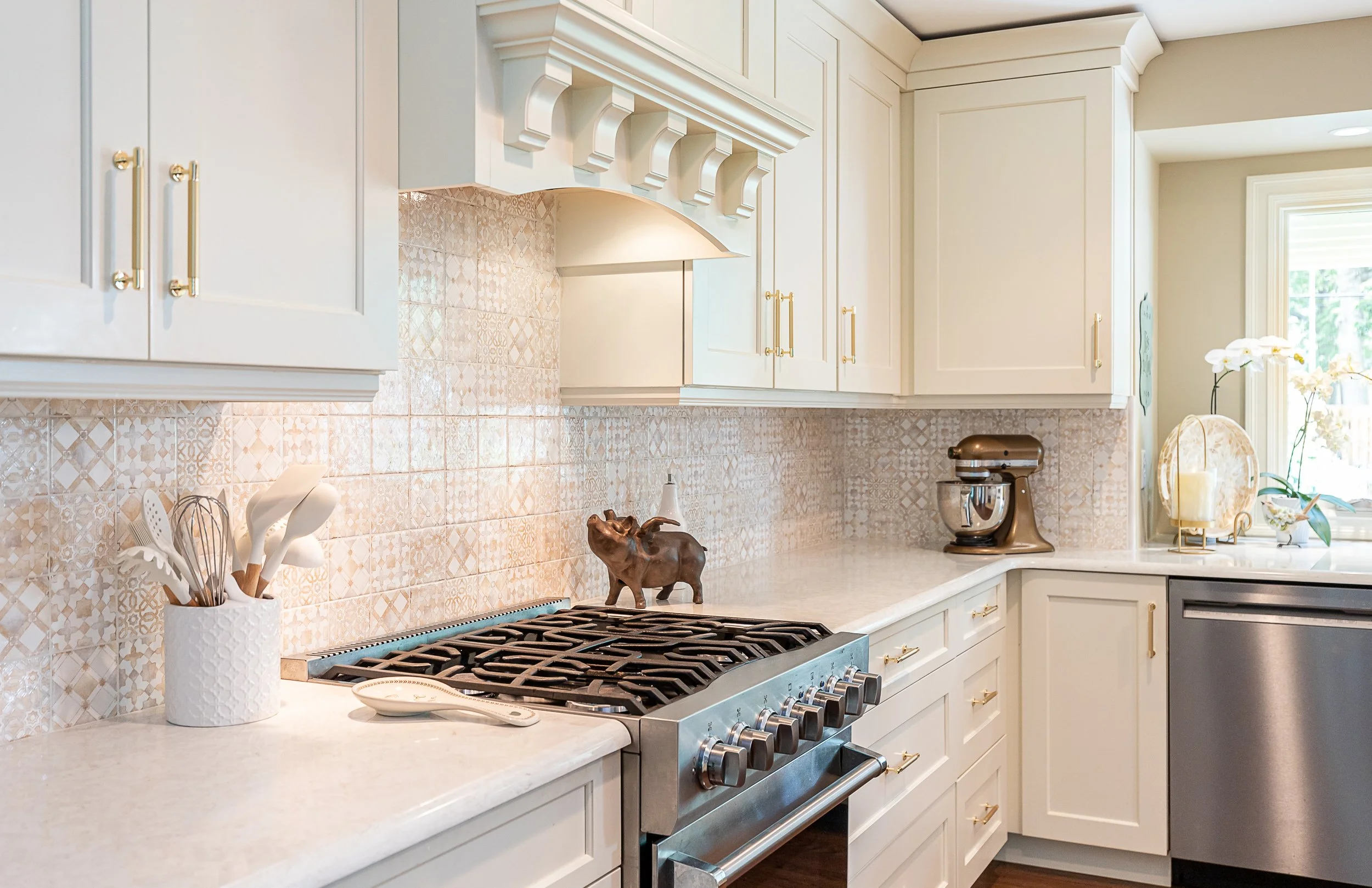 A modern beige kitchen with beige tiles, granite countertops, gold hardware, a gas stove, and decorative items including a brown pig figurine, a stand mixer, and orchids near a window.