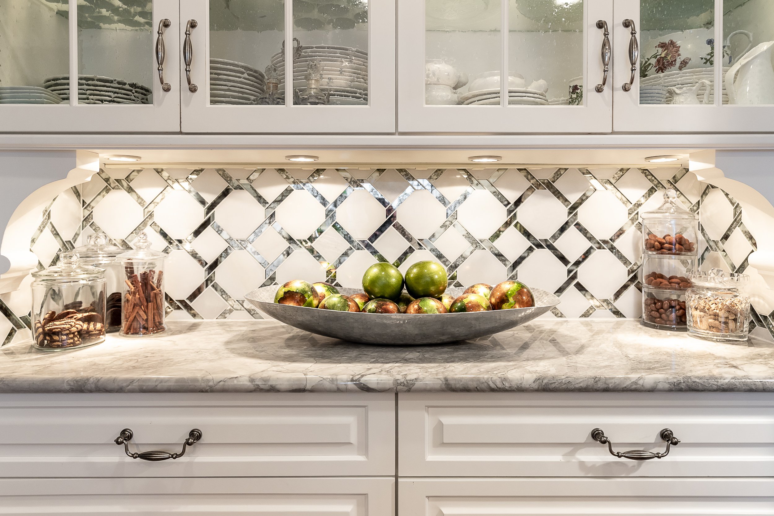 Decorative kitchen countertop with a large silver bowl of apples, glass jars of snacks, and a white cabinet with glass doors and china inside.