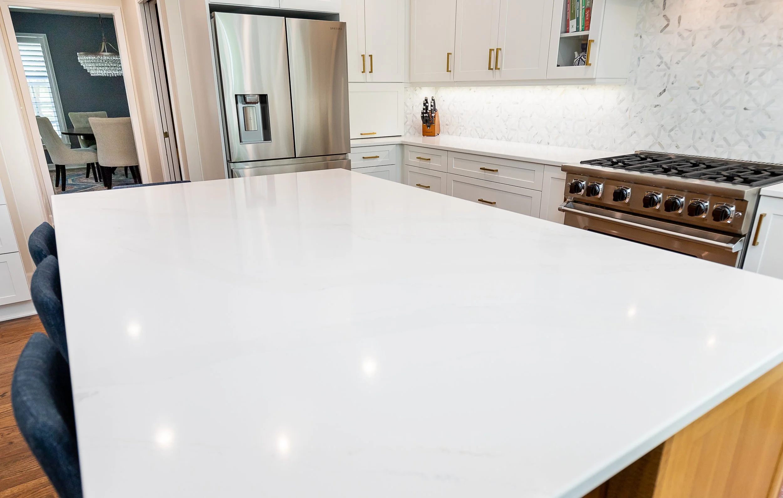 A modern white kitchen with a large white quartz countertop island, stainless steel refrigerator, and white cabinets with gold handles. Part of a dining room with a chandelier is visible through an open doorway.