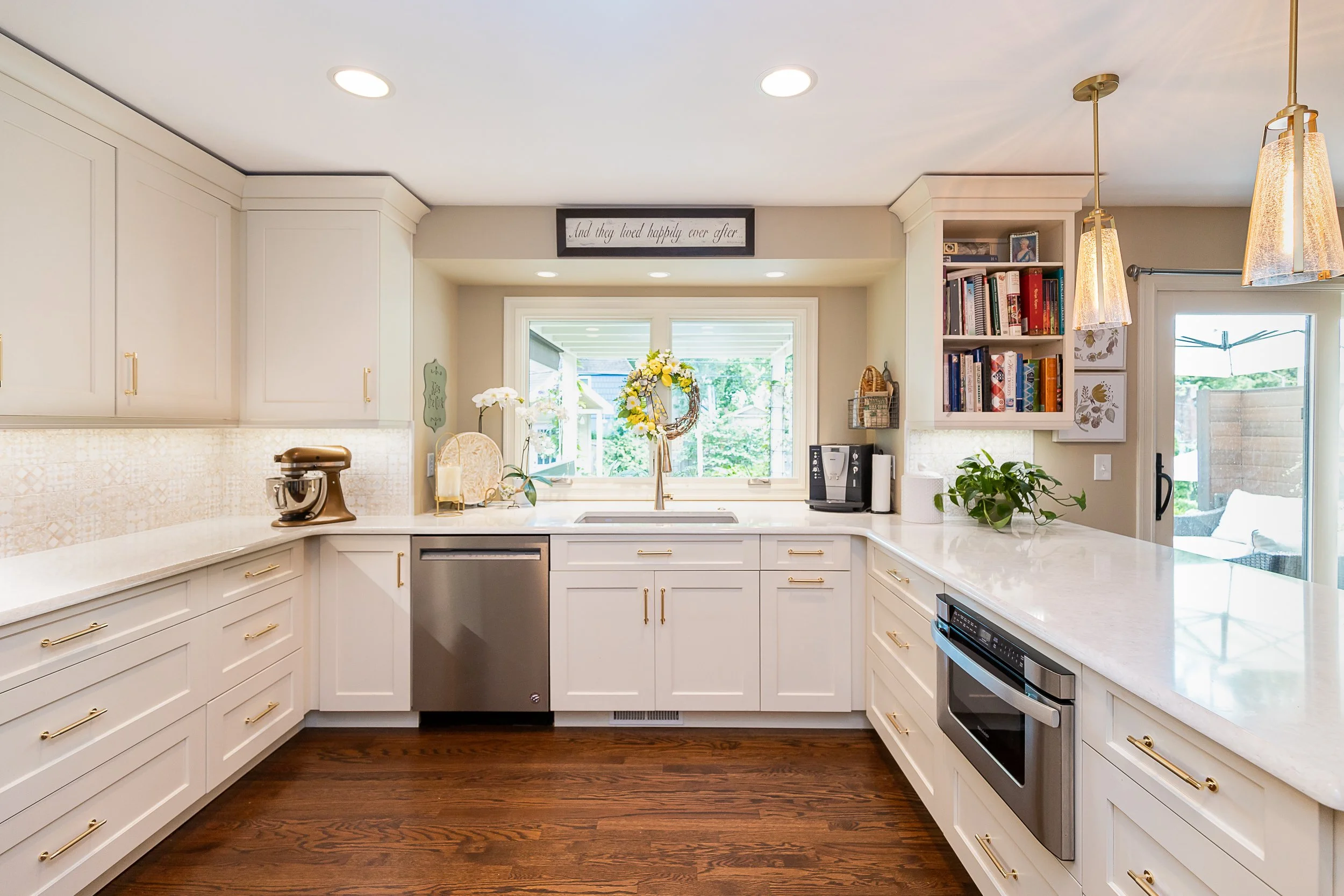 Bright kitchen with beige cabinets, gold hardware, a window with a view of greenery, and hardwood flooring.
