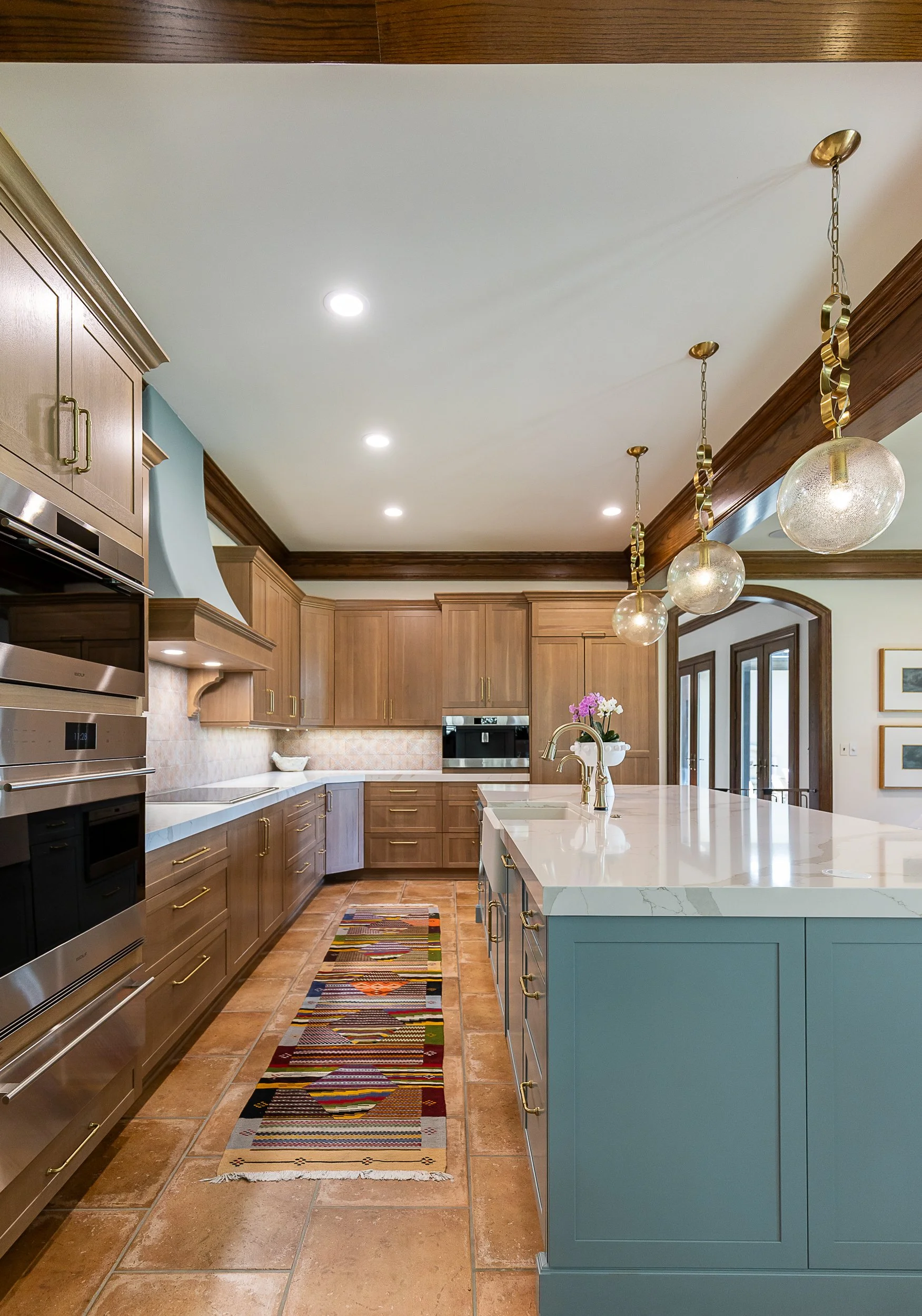 A spacious kitchen with wooden cabinetry, a white marble-looking island, pendant lights, and a colorful rug on the tiled floor.