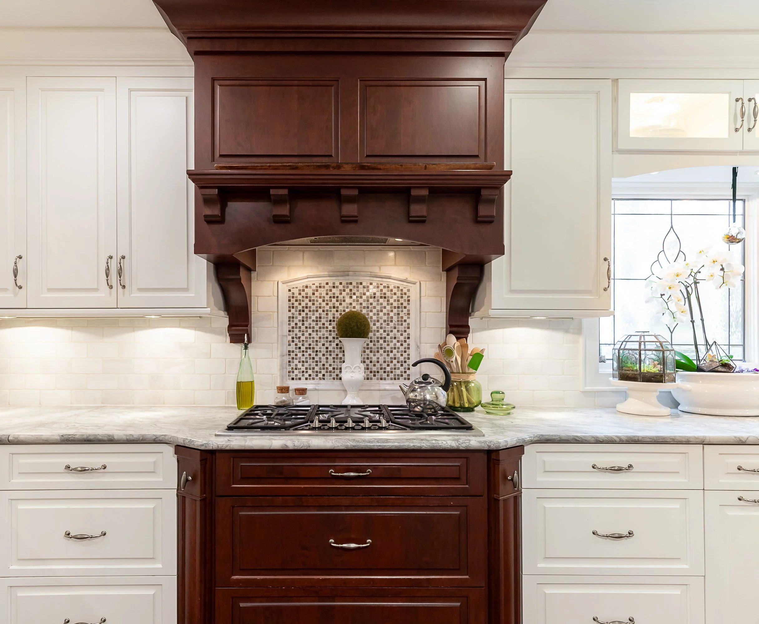 Kitchen with white cabinets, quartz countertops, and a dark wood range hood over a stove, with decorative plants and kitchen utensils on the counter.