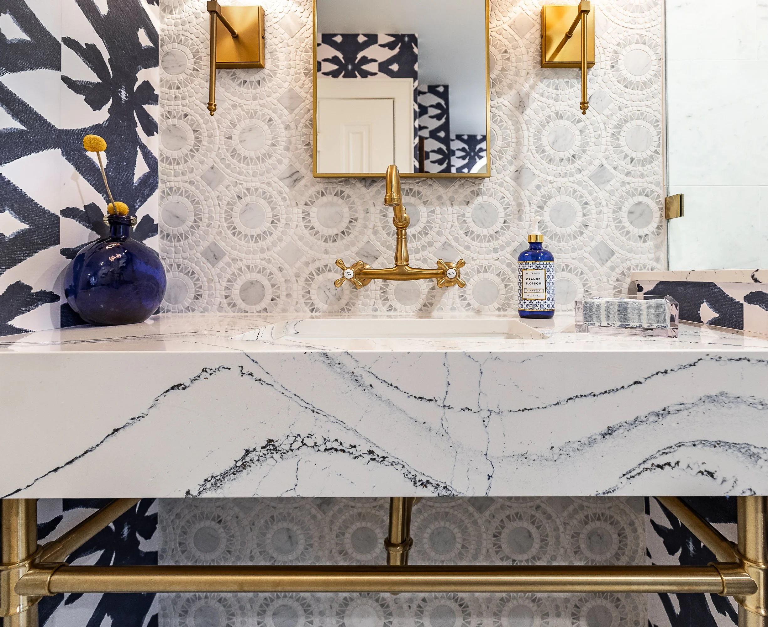 A stylish bathroom vanity with a white marble-looking console, gold fixtures, a blue vase with yellow-tipped flowers, a patterned tile backsplash, a mirror, and a blue bottle of hand soap.