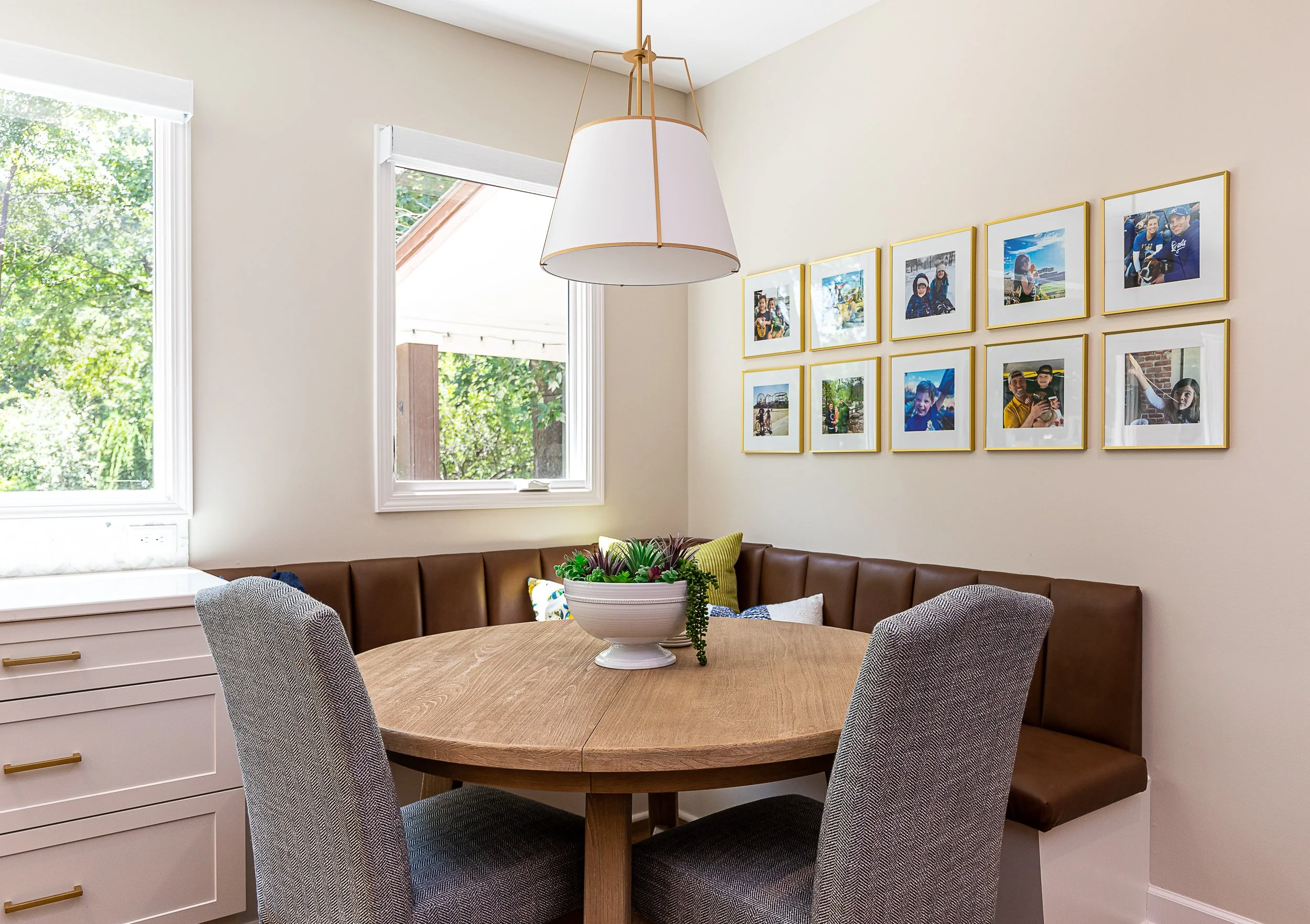 Dining nook with round wooden table, gray upholstered chairs, and a brown leather benchtop seating area, with a large white pendant light and a collage of framed photos on the cream-colored wall. Two windows with white frames and blinds show greenery