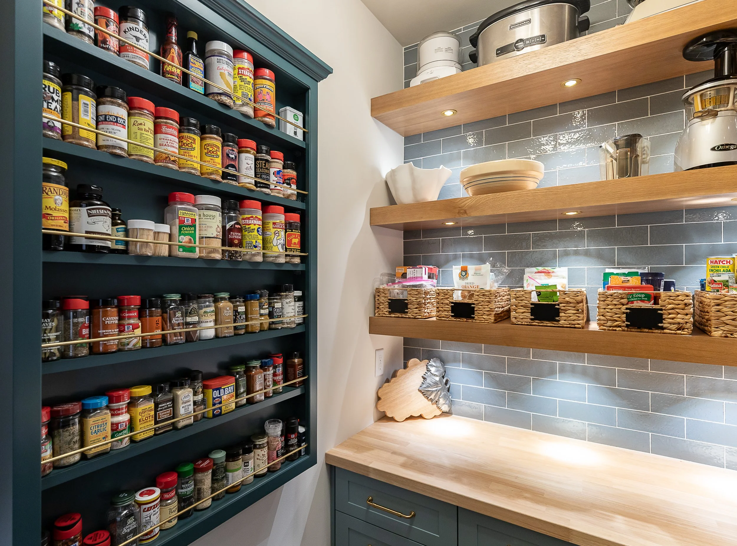 A kitchen pantry with open wooden shelves, bowls, and appliances, next to a blue spice cabinet filled with various seasonings.