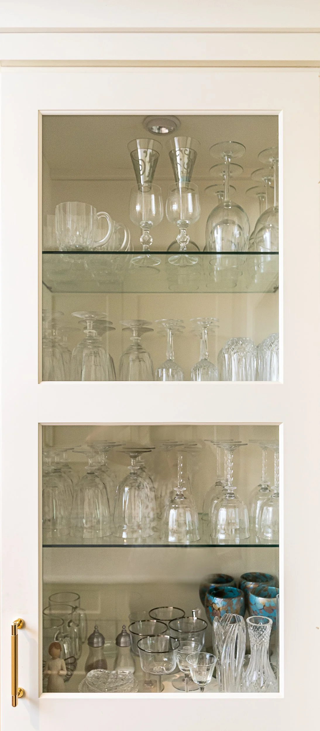 A beige kitchen cabinet with glass doors, filled with various glassware including wine glasses, champagne flutes, cups, and decorative bowls.
