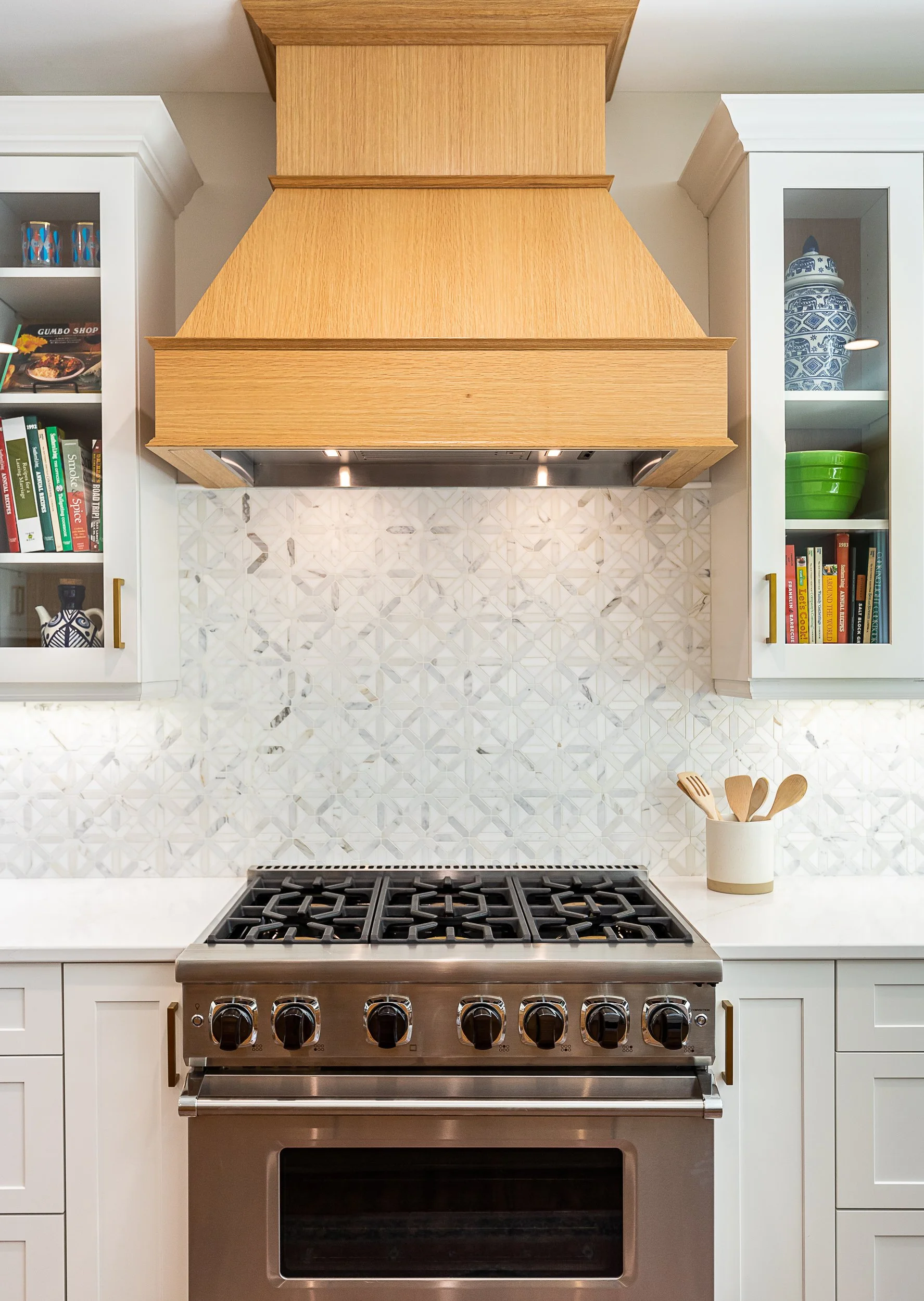 Modern kitchen with white cabinets, stainless steel gas stove, patterned tile backsplash, light wood range hood, and open shelves with cookbooks and decorative items.