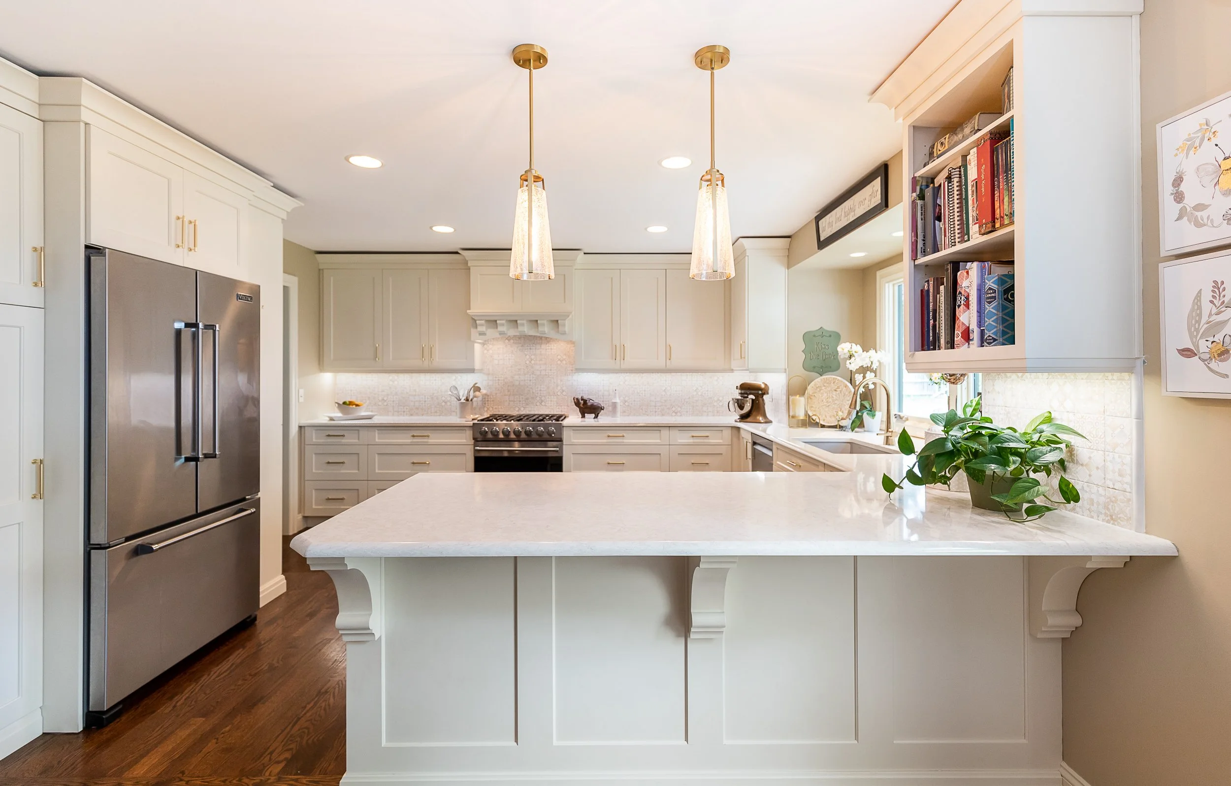 Modern kitchen with beige cabinetry, stainless steel refrigerator, and island with pendant lights, gold hardware, and decorative plants.