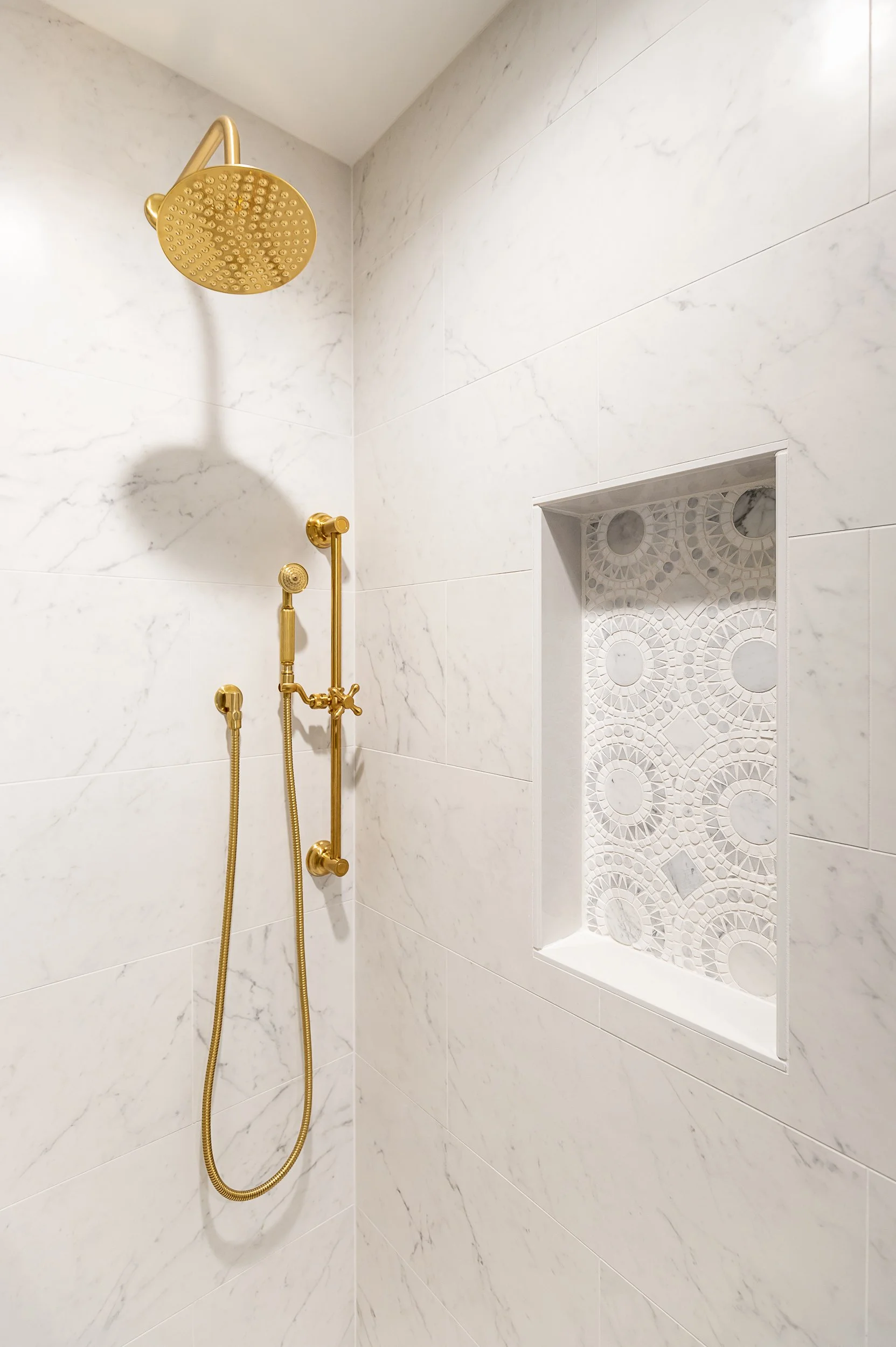 Shower with white marble-looking tiles, gold showerhead, and a built-in decorative niche with mosaic tiles.