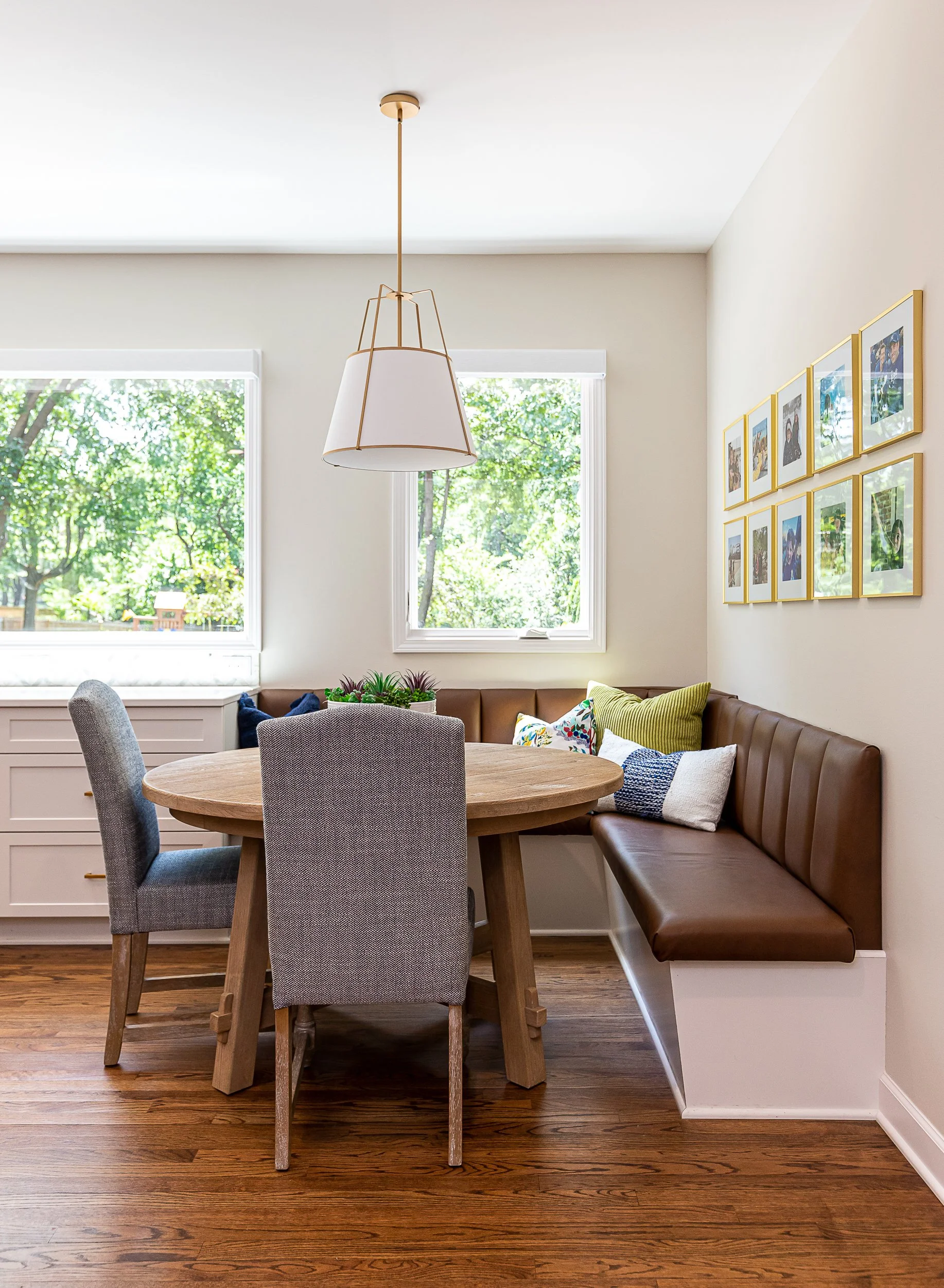 Kitchen nook with round wooden table, gray upholstered chairs, and a brown leather bench with colorful pillows, two large windows with greenery outside, framed photographs on the wall, and a modern white and gold pendant light.