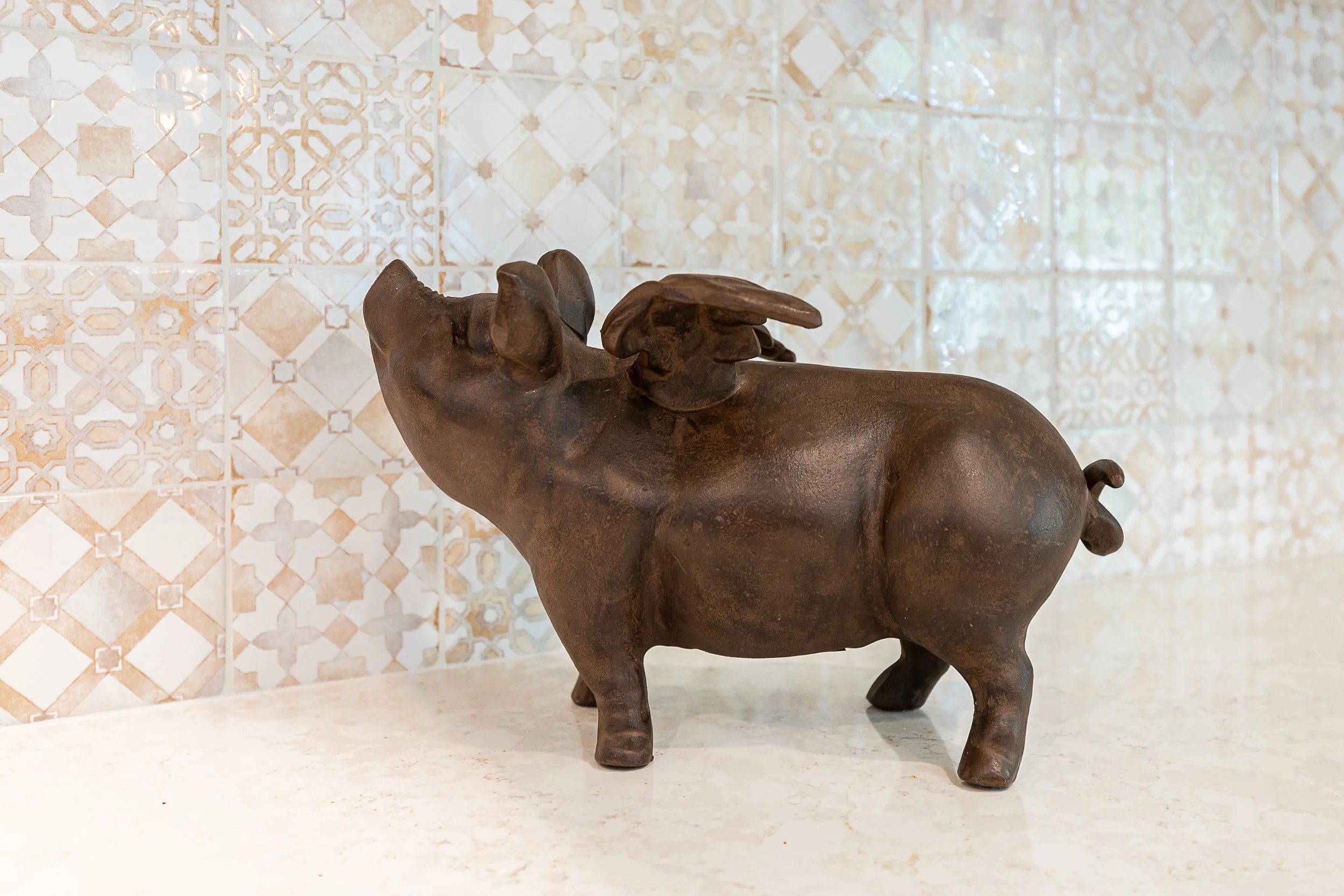 Bronze sculpture of a pig with a small bird on its back, placed on a quartz surface against a patterned tiled wall.