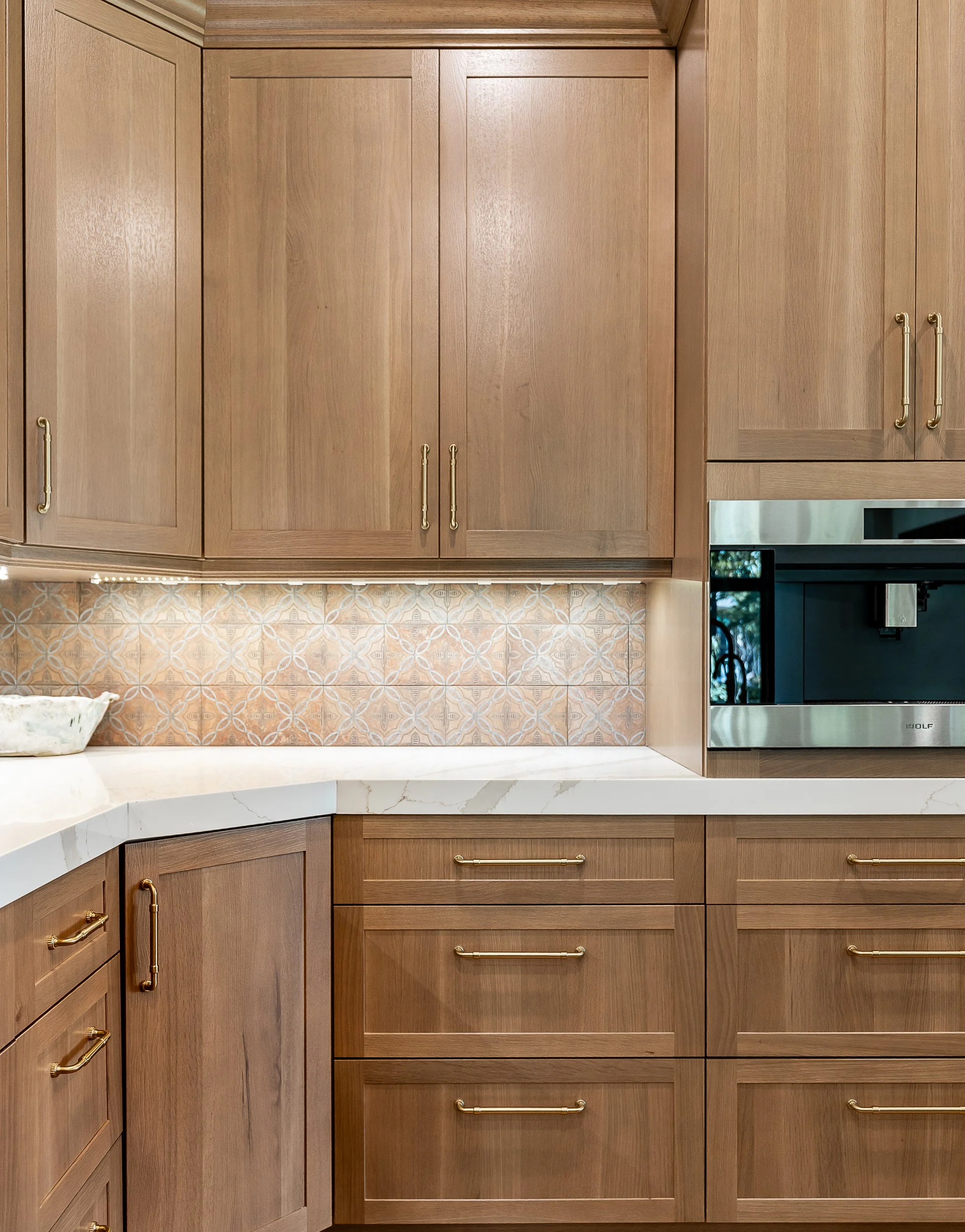 Kitchen cabinets made of light wood with gold handles above a white marble-looking countertop. A built-in stainless steel oven is partially visible on the right side, and decorative patterned tiles are behind the counter.