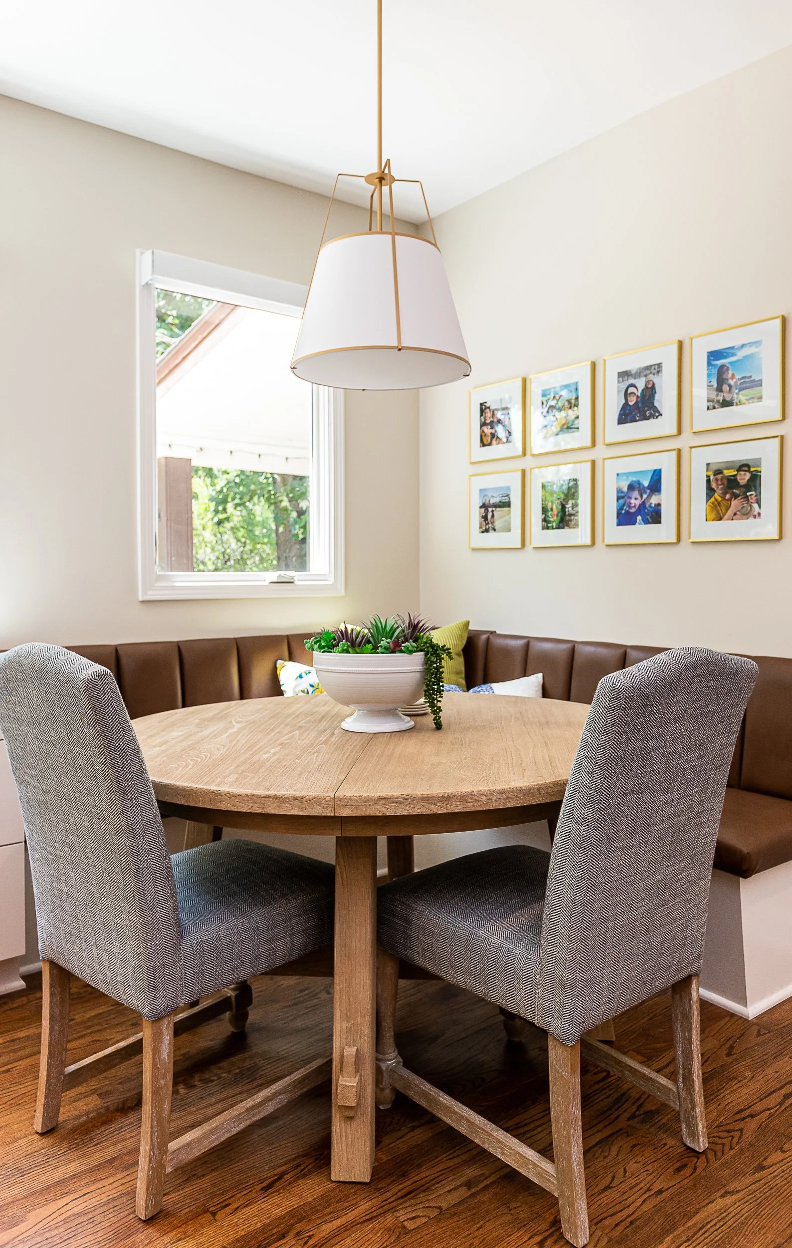 A cozy dining nook with a round wooden table, four upholstered chairs, and a built-in bench with cushions, illuminated by a modern pendant light. A window with a view of greenery and a wall decorated with framed photographs.