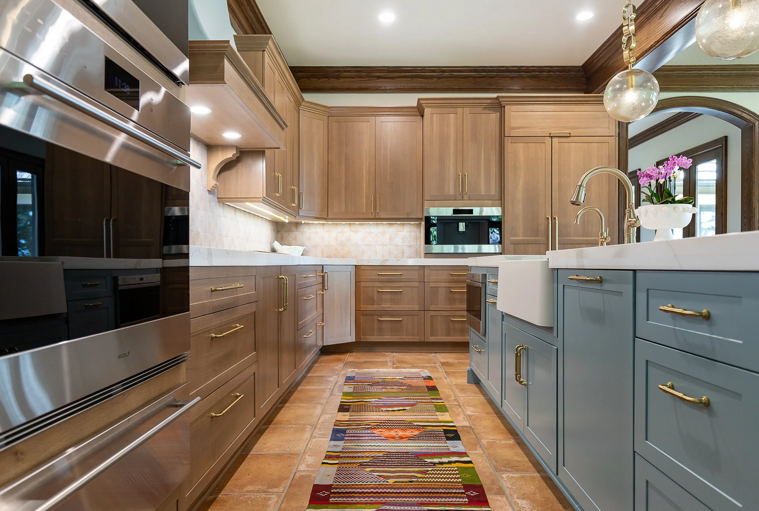A modern kitchen with wooden cabinets, a colorful striped rug on the floor, a kitchen island with a white countertop and blue cabinets, a gold faucet, purple flowers in a white pot, and a stainless steel oven, with a ceiling light and natural light coming through the window.