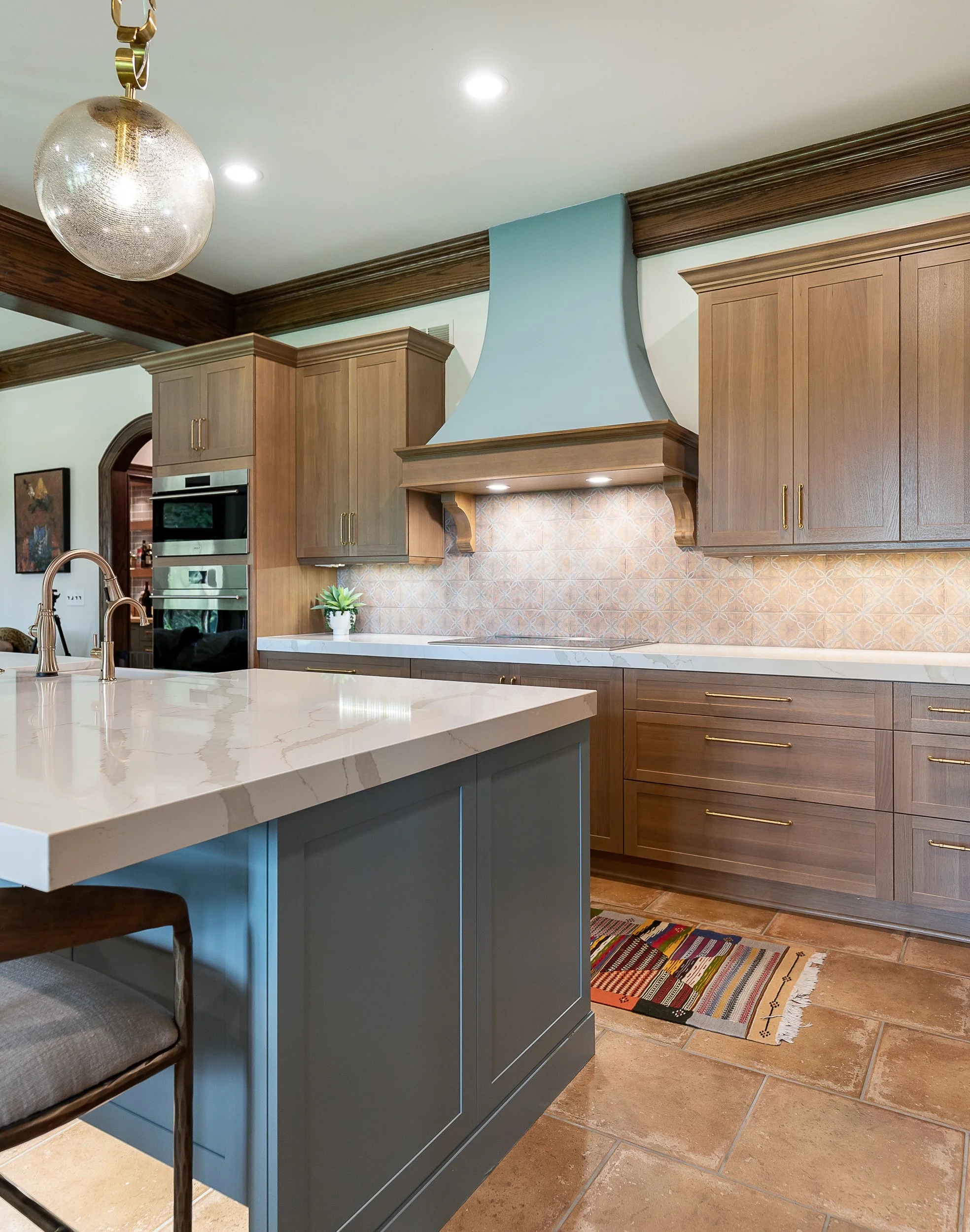 A kitchen with wooden cabinets and a white countertop island. There is a hanging glass light fixture, a small potted plant, and colorful rugs on the tiled floor.