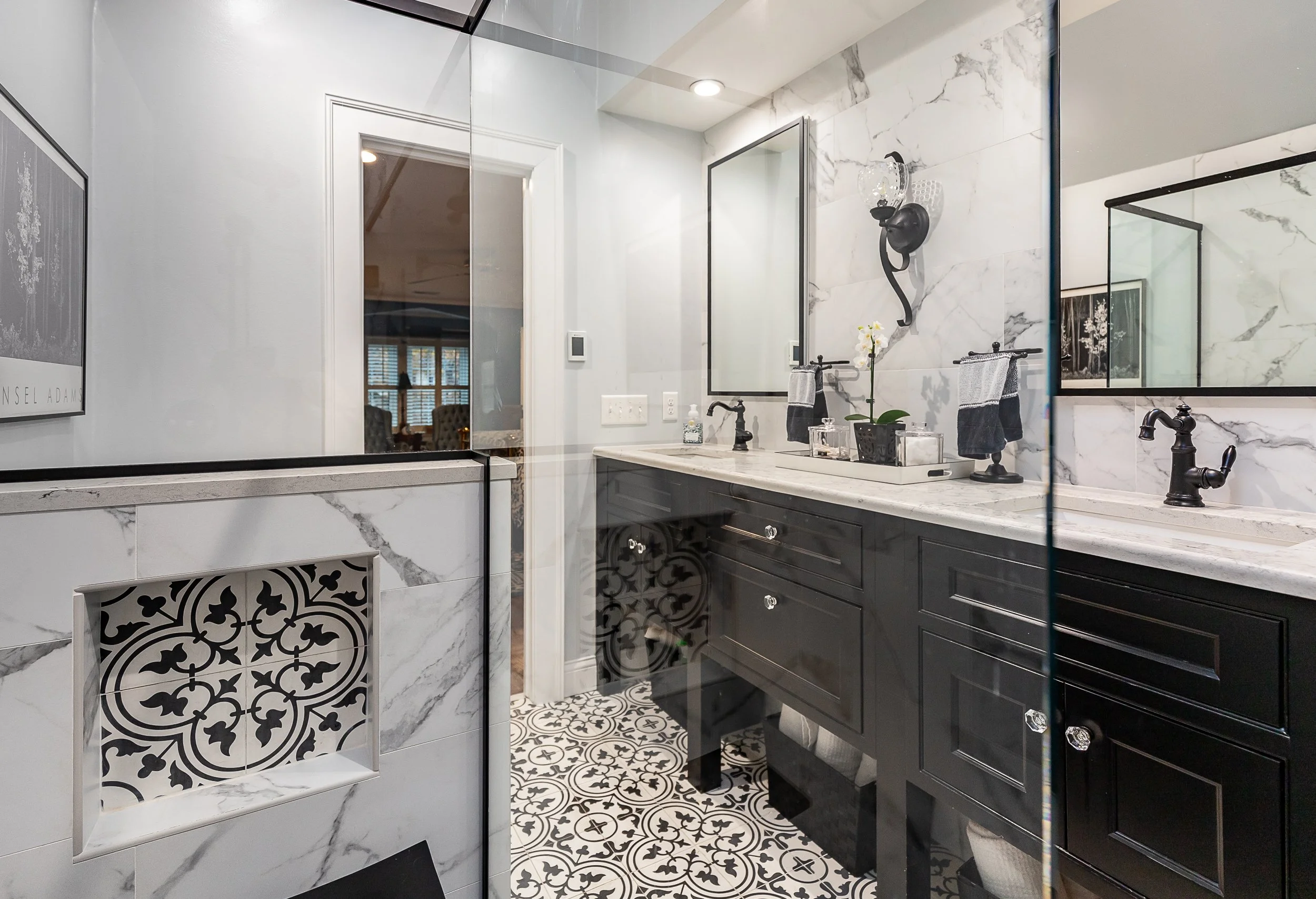 A modern bathroom with white marble-like tile walls, black fixtures, and a patterned tile floor. Features a black vanity with a white stone countertop, two mirrors, and decorative items.