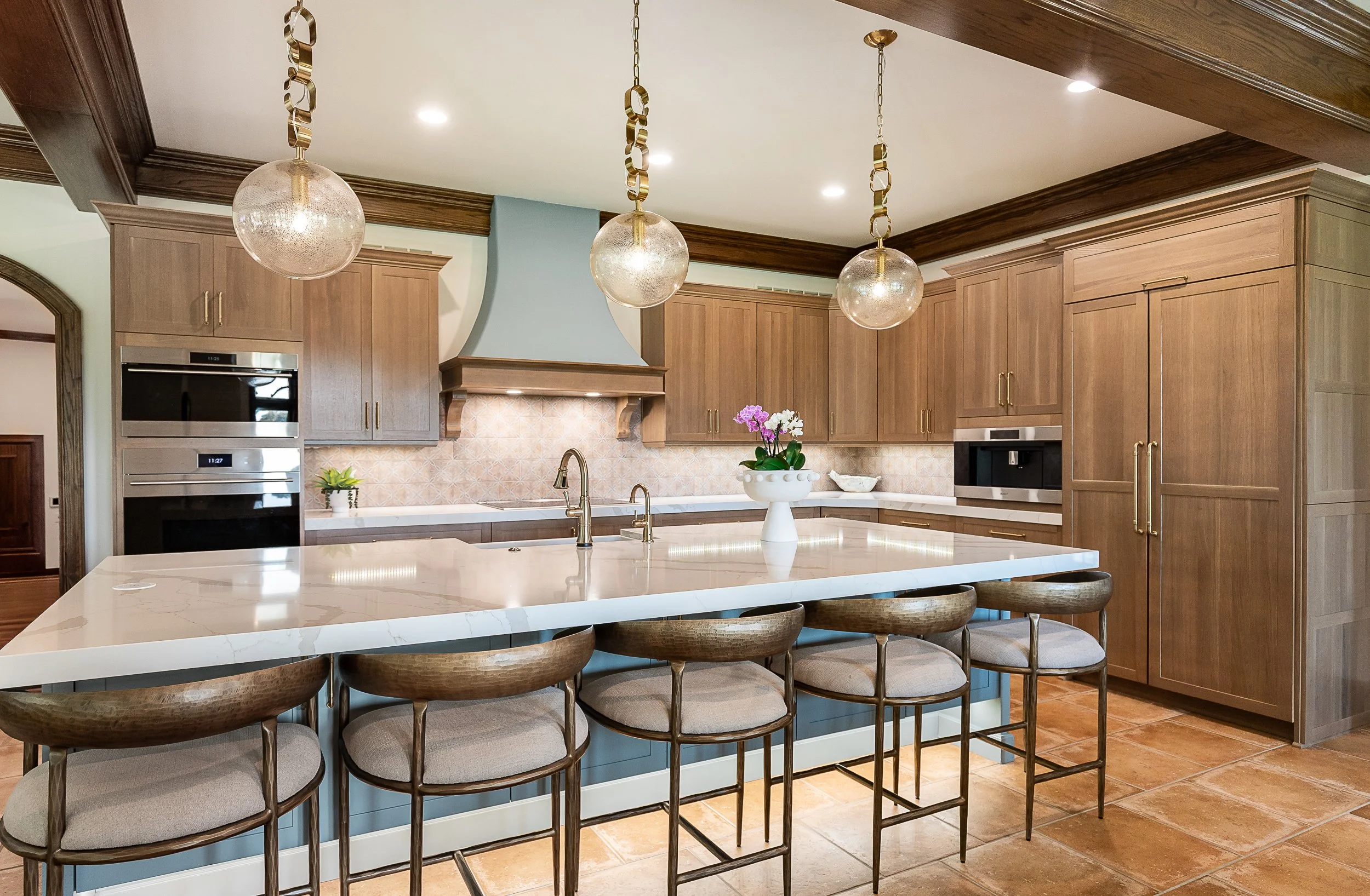 Kitchen with wooden cabinets, a white marble island with bar stools, pendant lights hanging from the ceiling, and a flower vase on the island.