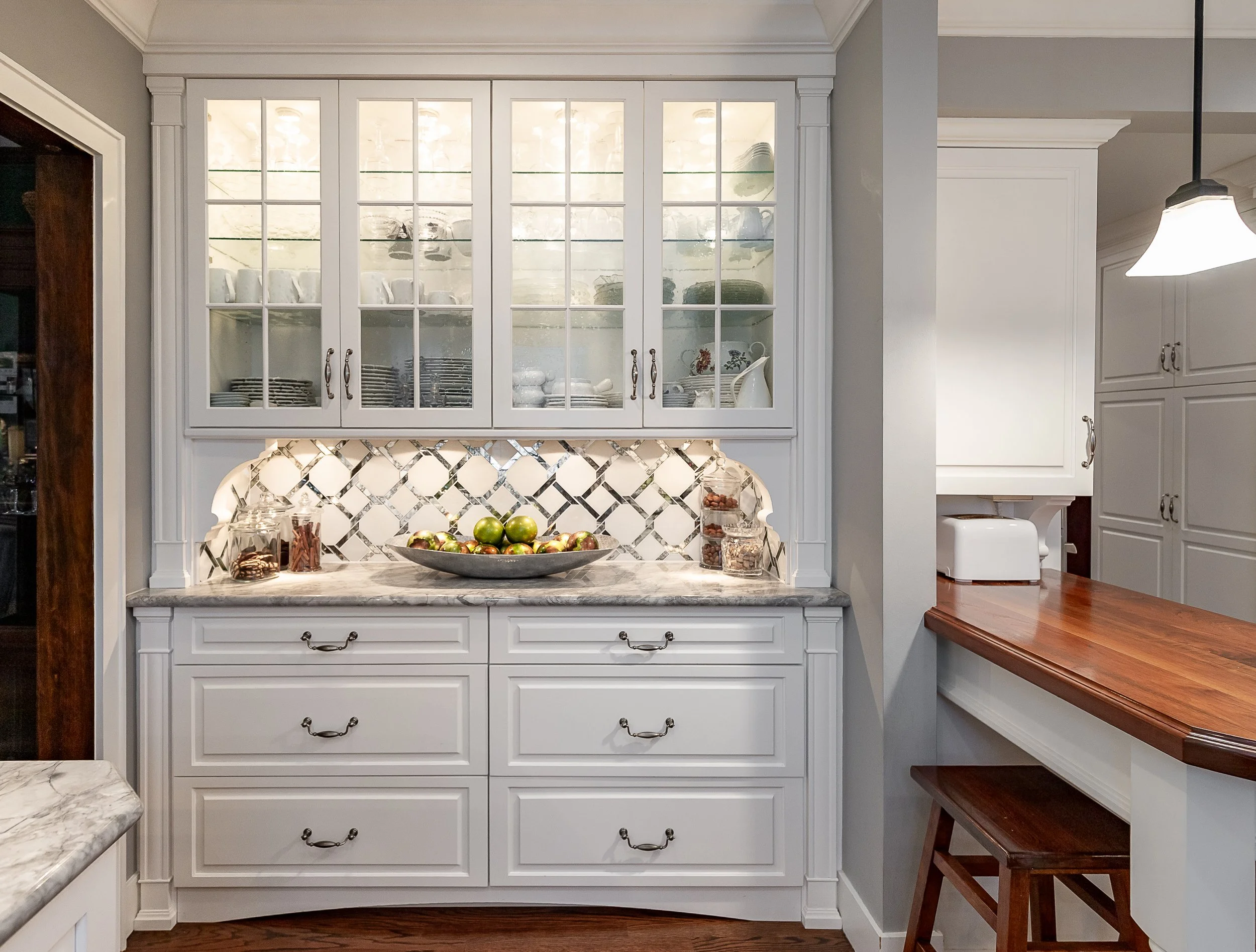 White kitchen cabinet with glass doors displaying dishes, a quartz countertop with a bowl of apples, jars, and a backsplash with a diamond pattern, adjacent to a wooden countertop and stool.