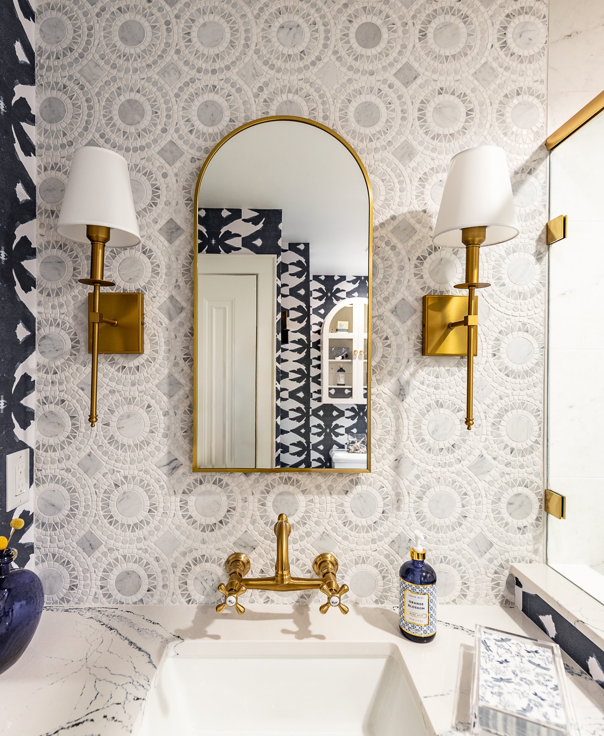 A bathroom vanity with a white marble countertop, gold fixtures including a faucet and wall sconces. A mirror with a gold frame hangs above the sink, reflecting a door.