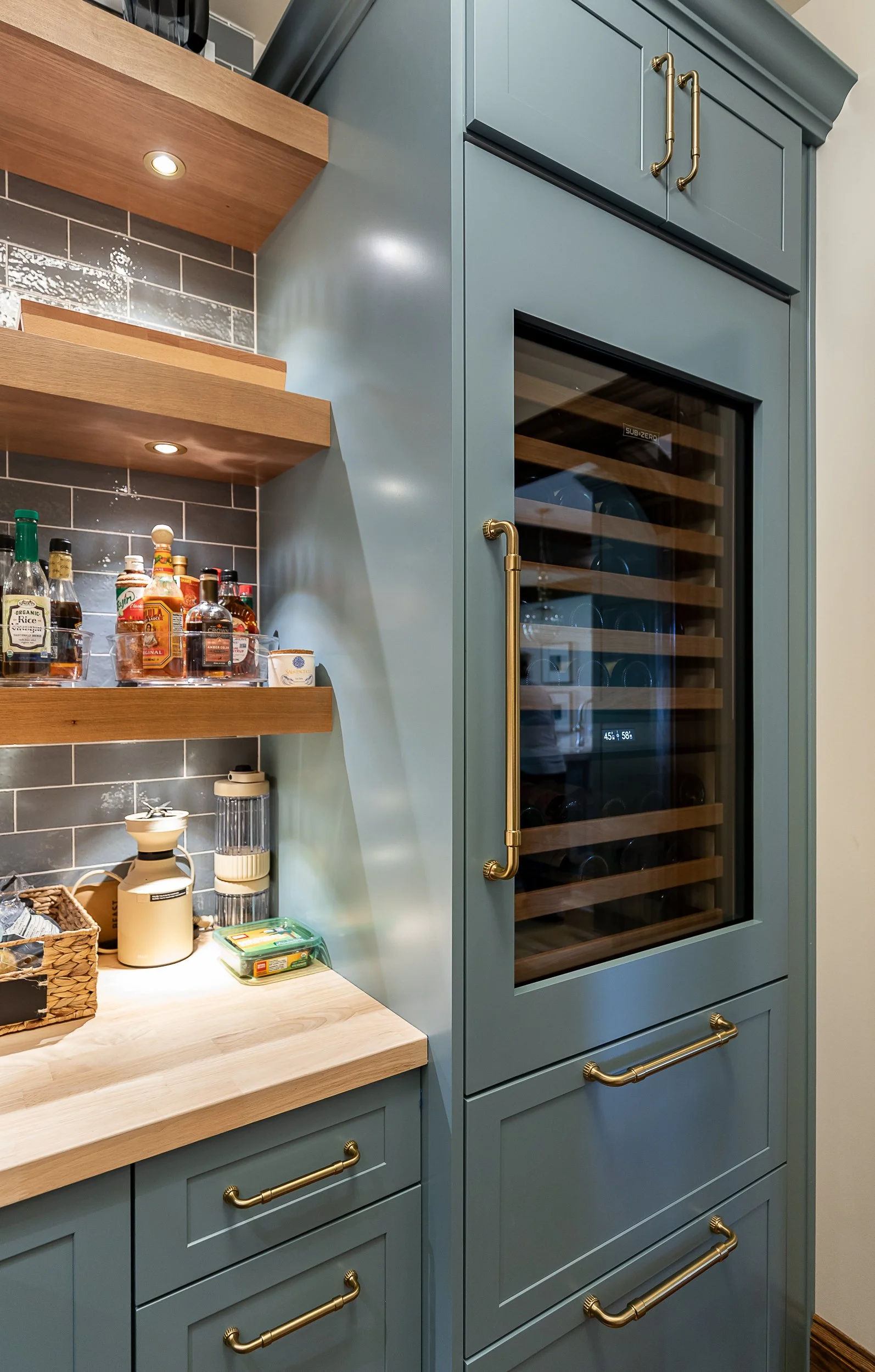 Kitchen with a built-in blue wine cooler with wooden shelves, gold handles, and a clear glass door, alongside a wooden countertop and open shelves with bottles, jars, and kitchen items.