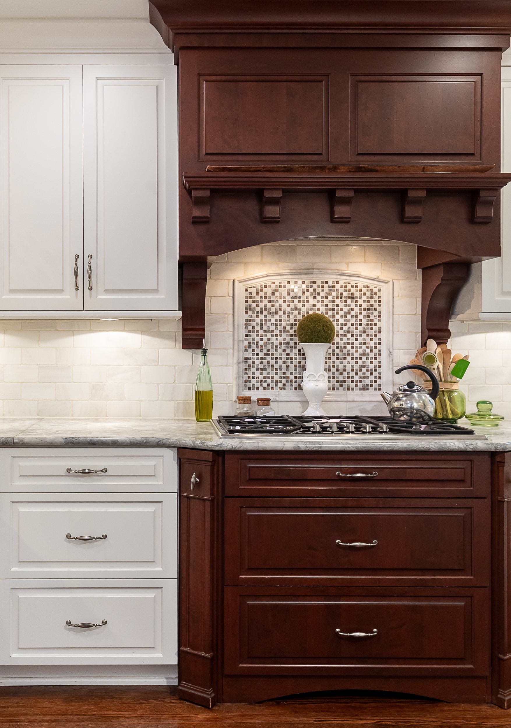 Kitchen with white and dark wood cabinets, a gas stove with a tile backsplash, a teapot, a vase with a round green plant, and kitchen utensils.