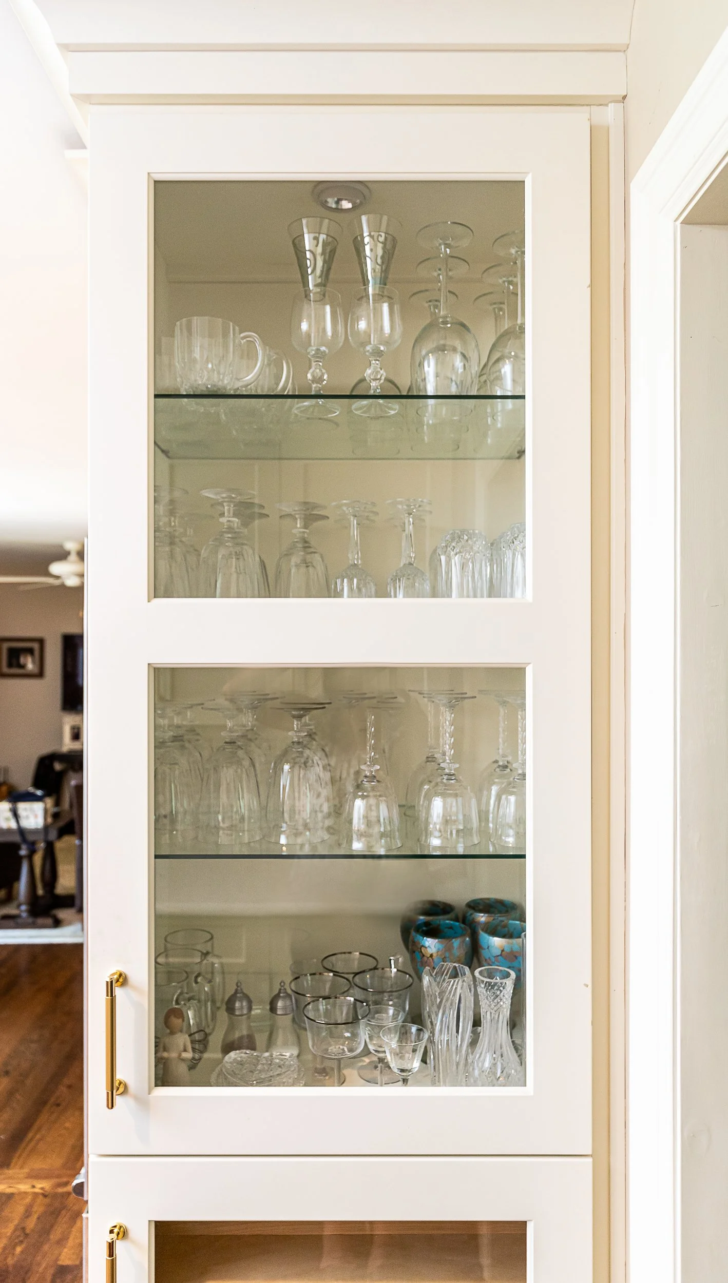 A beige glass cabinet with four shelves, displaying various glassware including wine glasses, champagne flutes, bowls, and decorative cups.