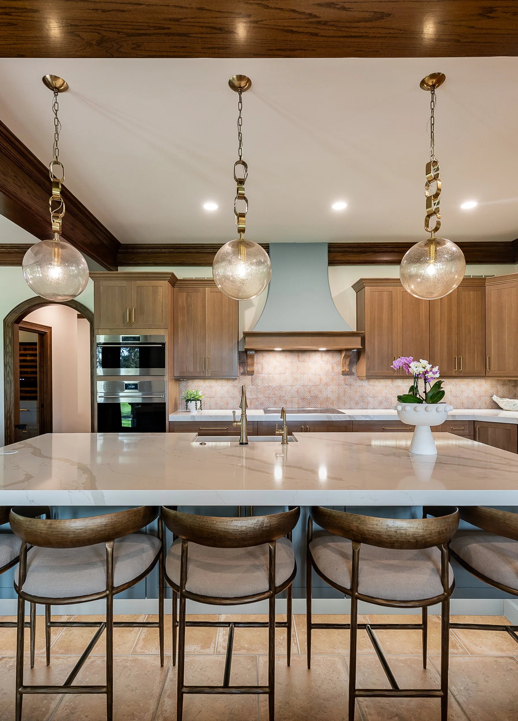 Modern kitchen with a large white marble island, wooden cabinets, beige tiled flooring, and three round pendant lights with glass globes hanging from brass chains.