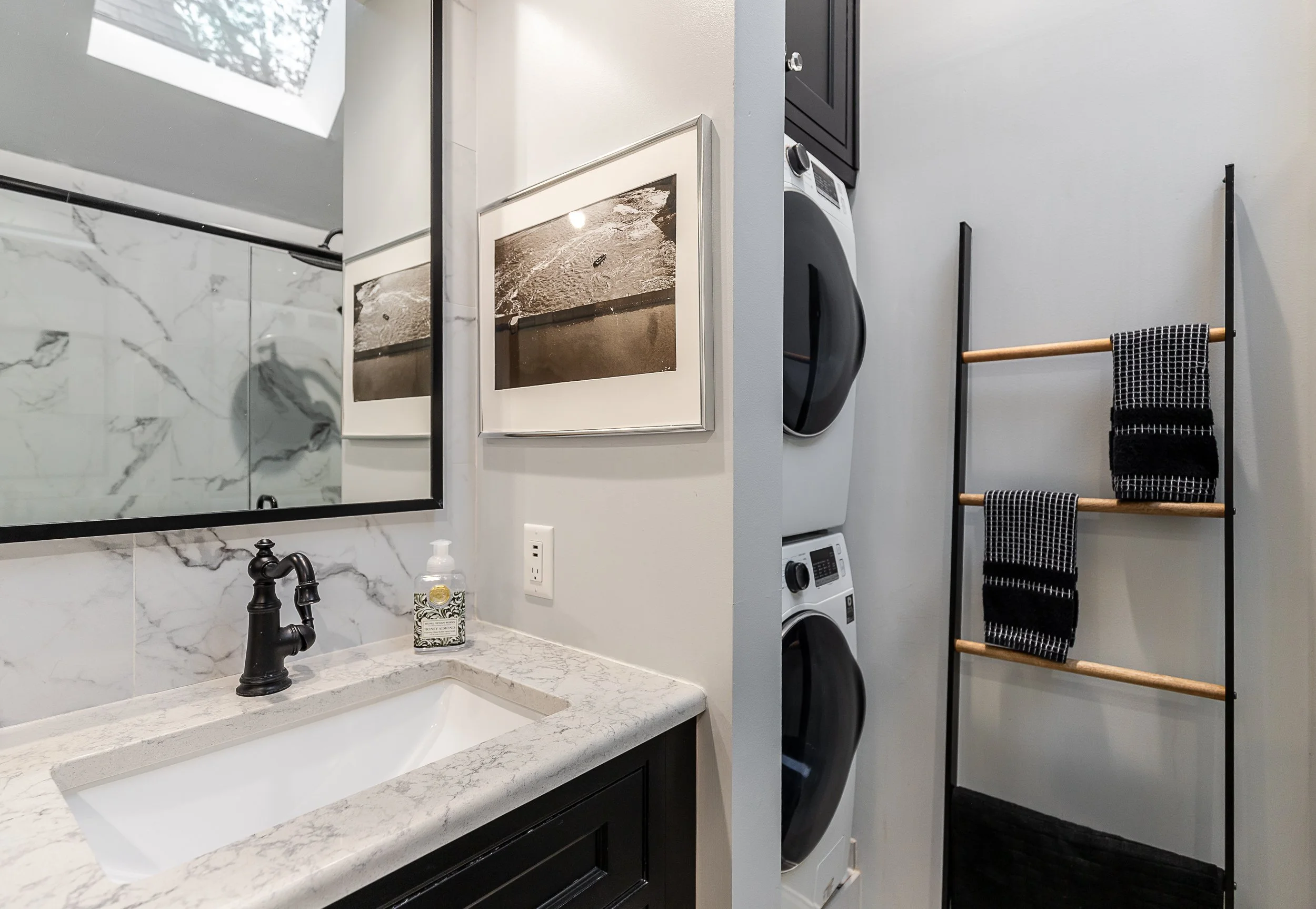 A bathroom with a marble-looking countertop, black faucet, framed black and white beach-themed photographs, a washer and dryer stacked in a closet, and a black ladder-style towel rack with two black towels.