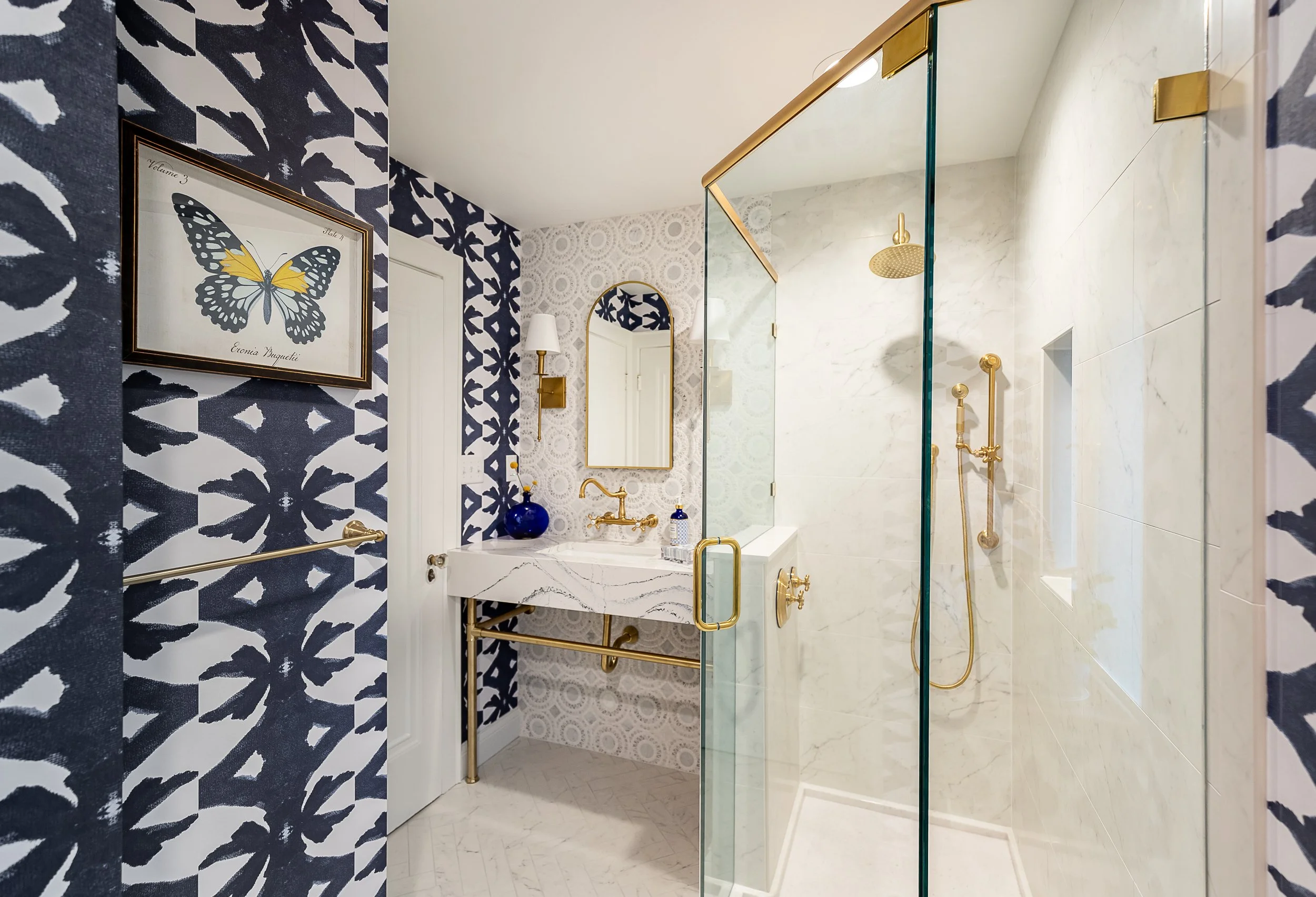 Modern bathroom with blue and white patterned wallpaper, white marble sink, gold fixtures, and a glass-enclosed shower with marble walls.