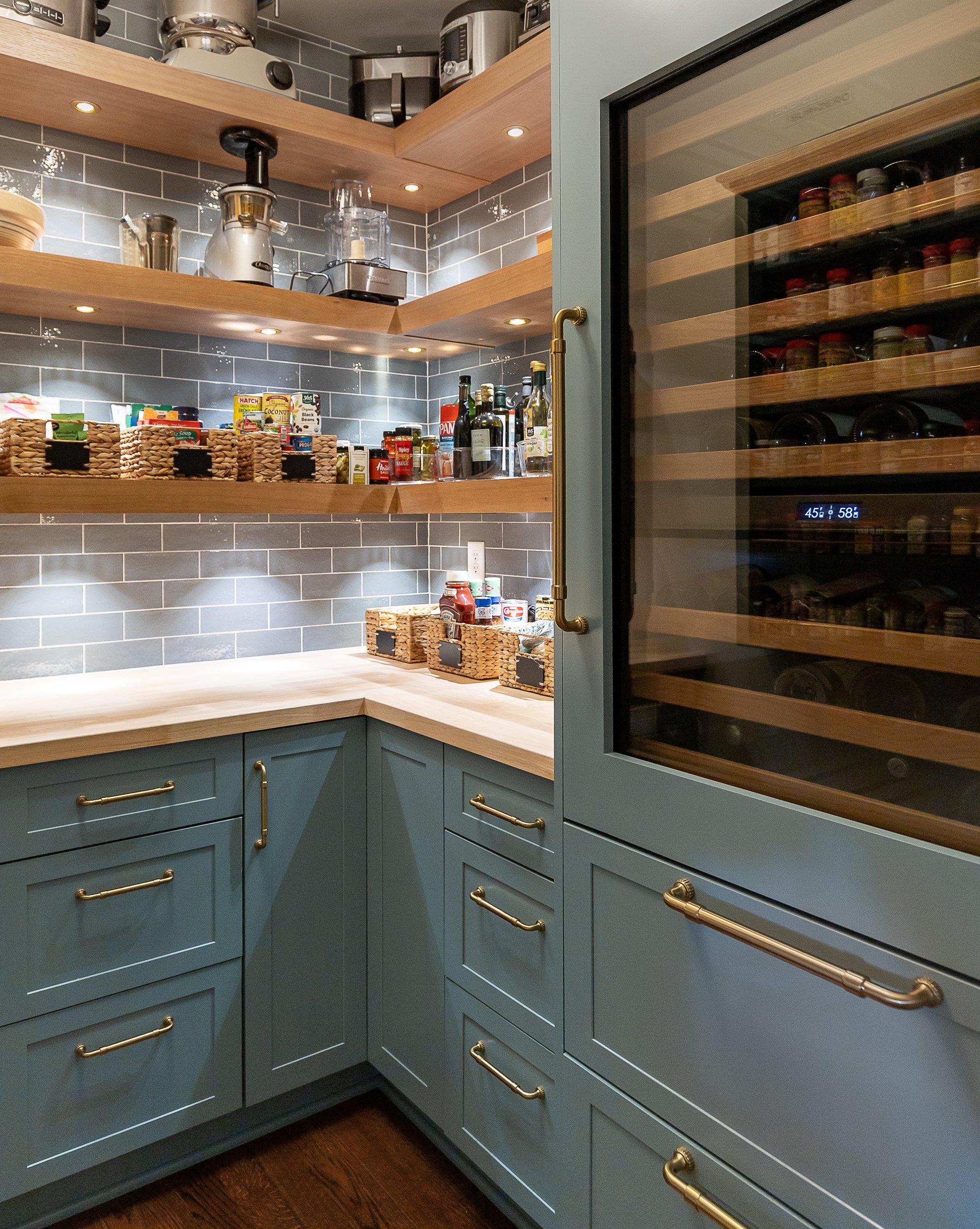 A kitchen corner with blue cabinets featuring gold handles, gray subway tile backsplash, wooden open shelves with kitchen appliances and pantry items, and a glass-front wine cooler.