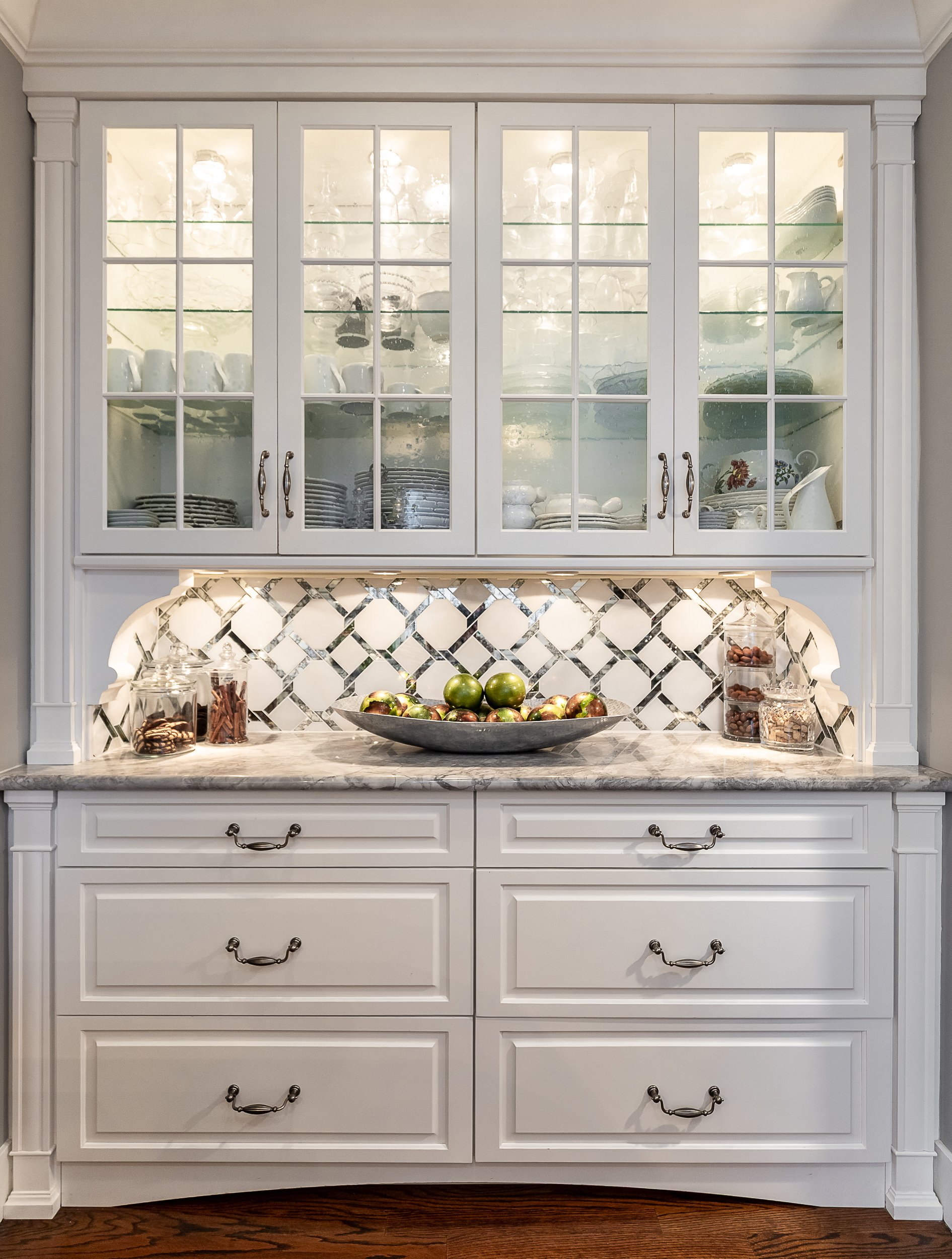 A white kitchen hutch with glass doors displaying dishes and glassware. The countertop below has a decorative tile backsplash and holds a large bowl of apples, glass jars of snacks, and a marble surface.