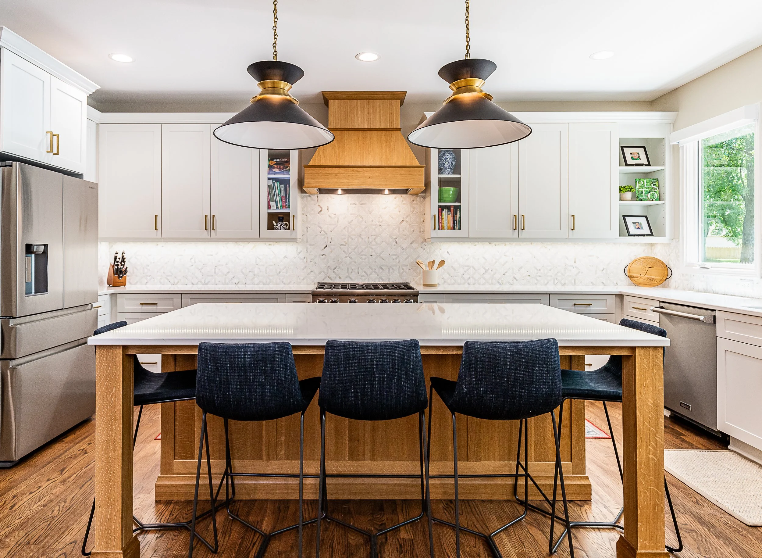 Modern kitchen with white cabinets, a large kitchen island with a white countertop, black chairs, a stainless steel refrigerator, a stove with a wooden range hood, and open shelves with books and decorations, illuminated by natural light from a window.