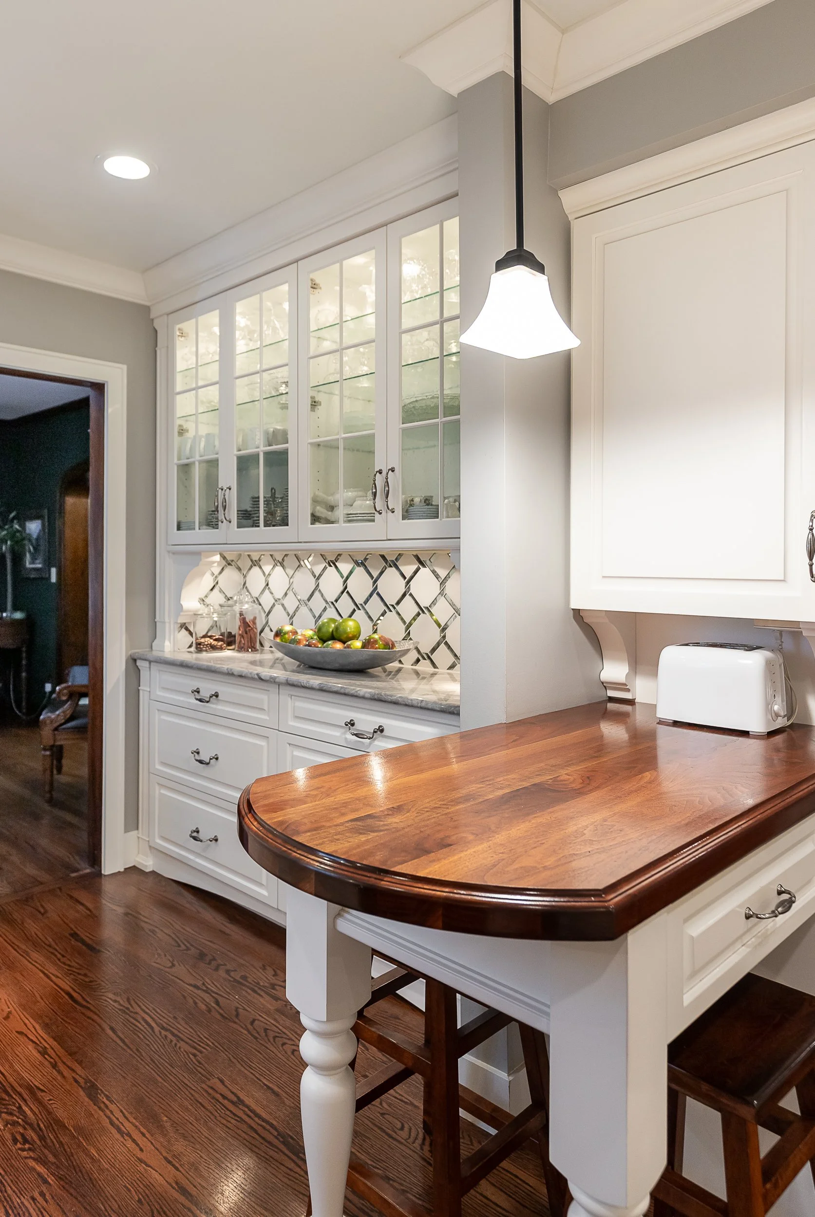 A kitchen with white cabinets, a wooden counter, and a hanging light. Glass-front upper cabinets and a quartz countertop are visible, with a bowl of apples and a glass jar on the counter.