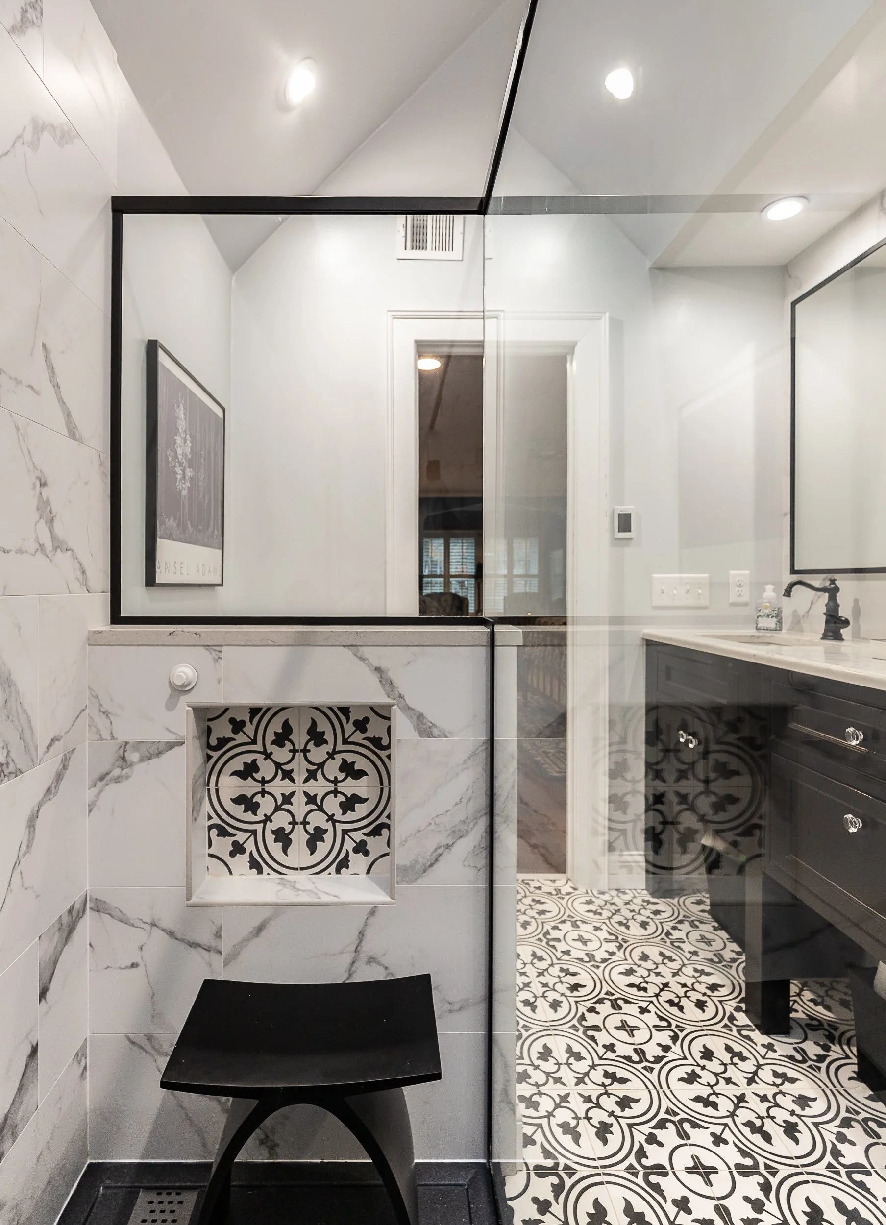 A bathroom with a glass partition, marble-looking wall tiles, a black stool, patterned black and white floor tiles, a black vanity with a white countertop, and a wall-mounted mirror.