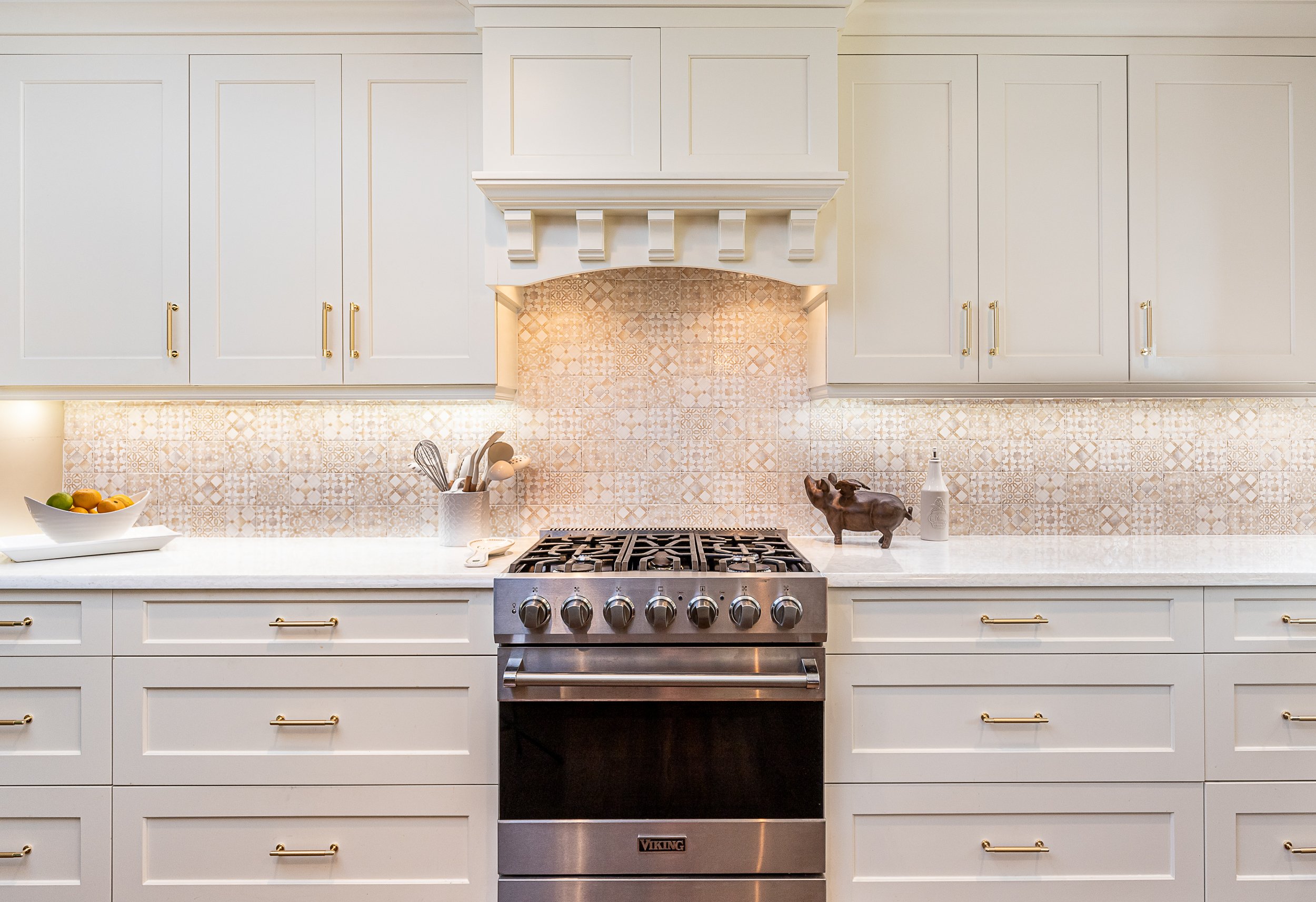 Kitchen with beige cabinets, beige patterned tile backsplash, stainless steel Viking oven, and decorative items including a ceramic pig and a white bottle on the countertop.