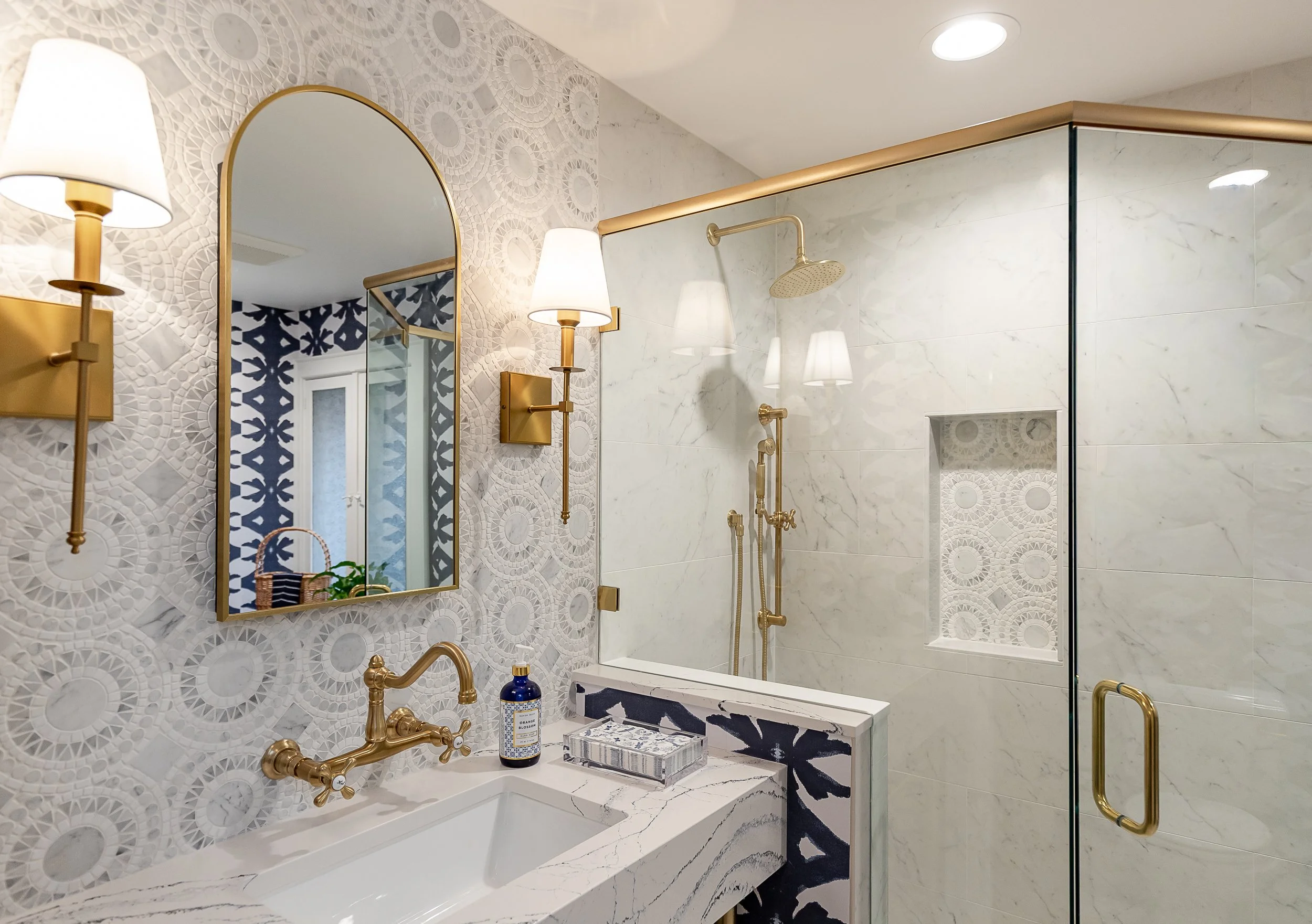 A modern bathroom featuring a white marble console with gold fixtures, a large mirror, wall-mounted gold lights, and a separate shower area with a glass door and gold fixtures.