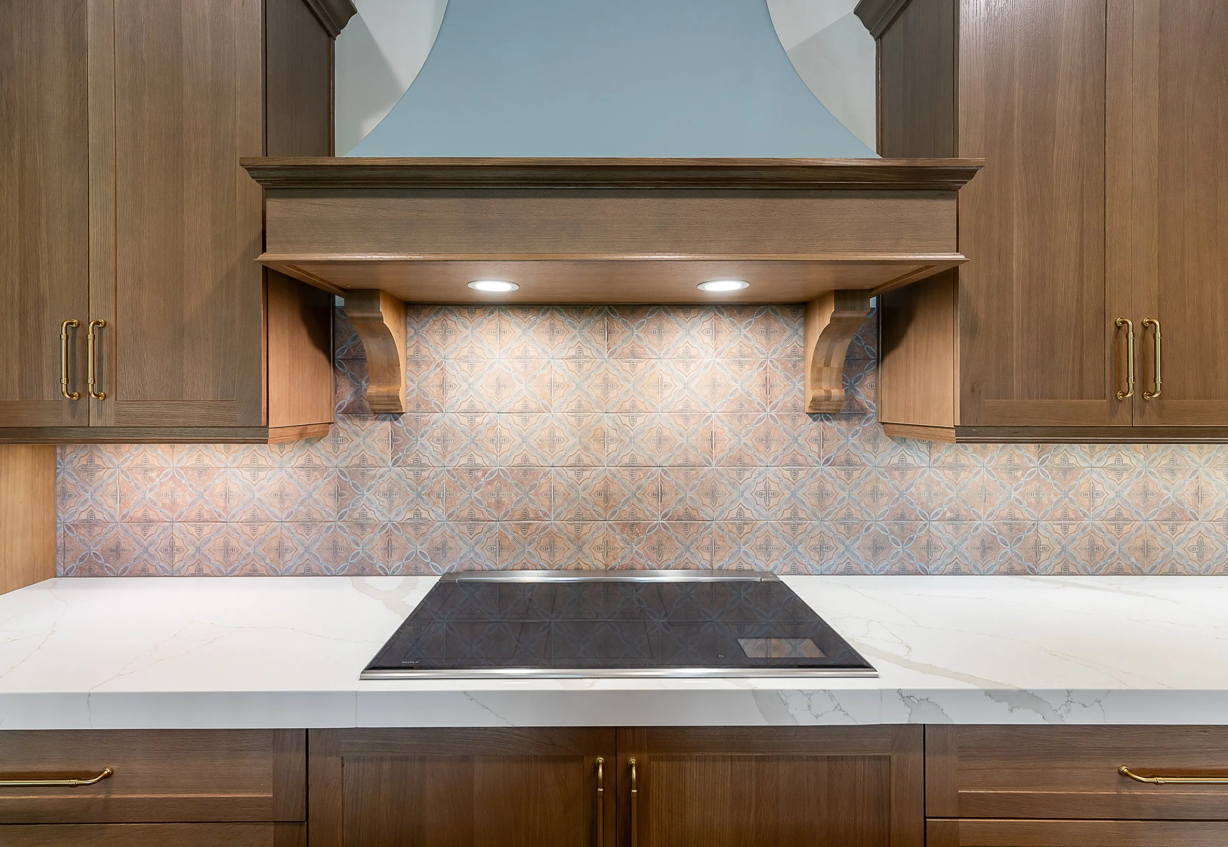 Close-up of a kitchen backsplash with patterned tiles, wooden upper cabinets, a black induction cooktop on a white marble countertop, and wooden lower cabinets with brass handles.