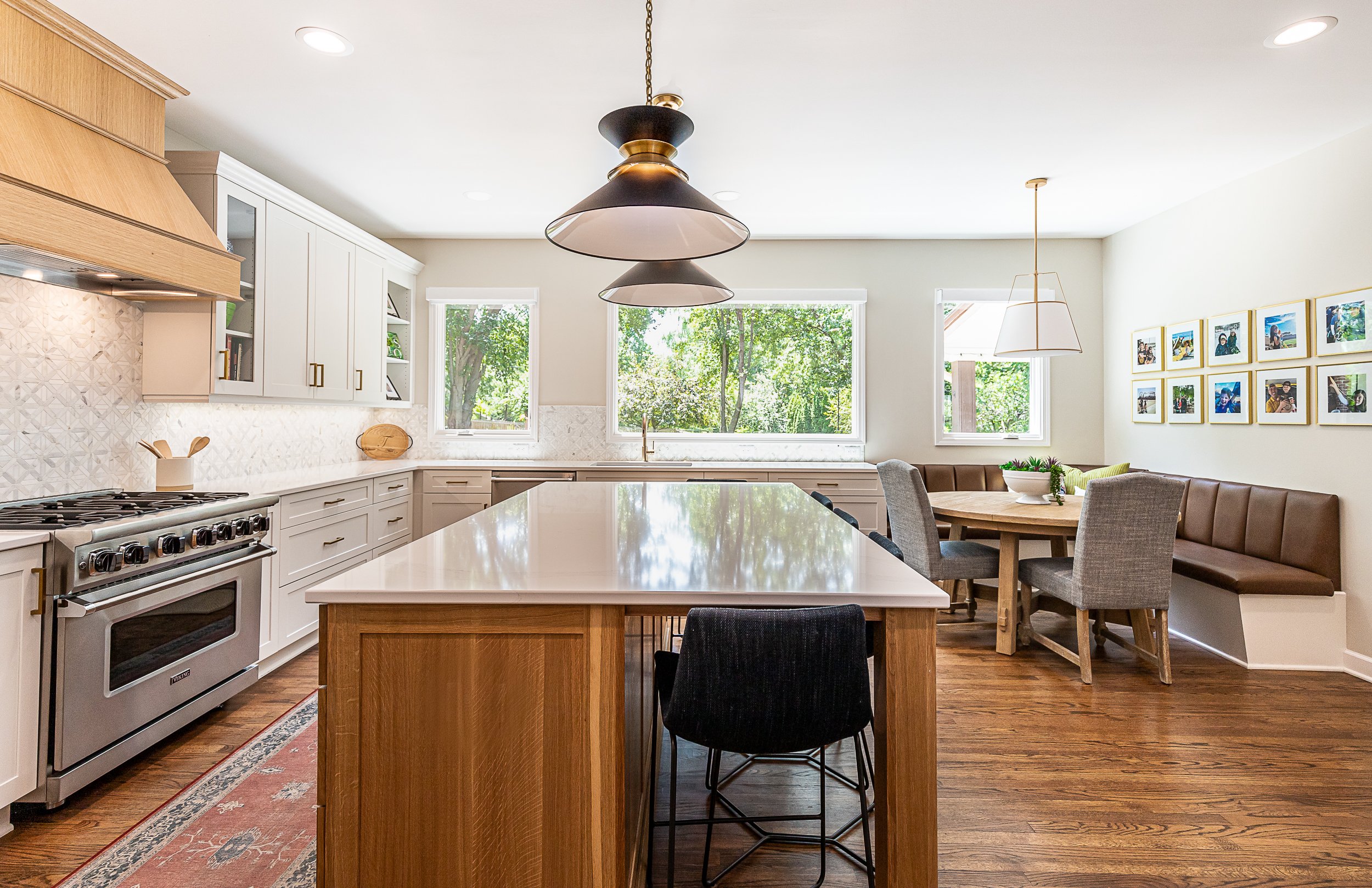 Bright, modern kitchen with white cabinetry, a large island with a white countertop, stainless steel oven, and a dining area with a round table, upholstered chairs, and framed photos on the wall.