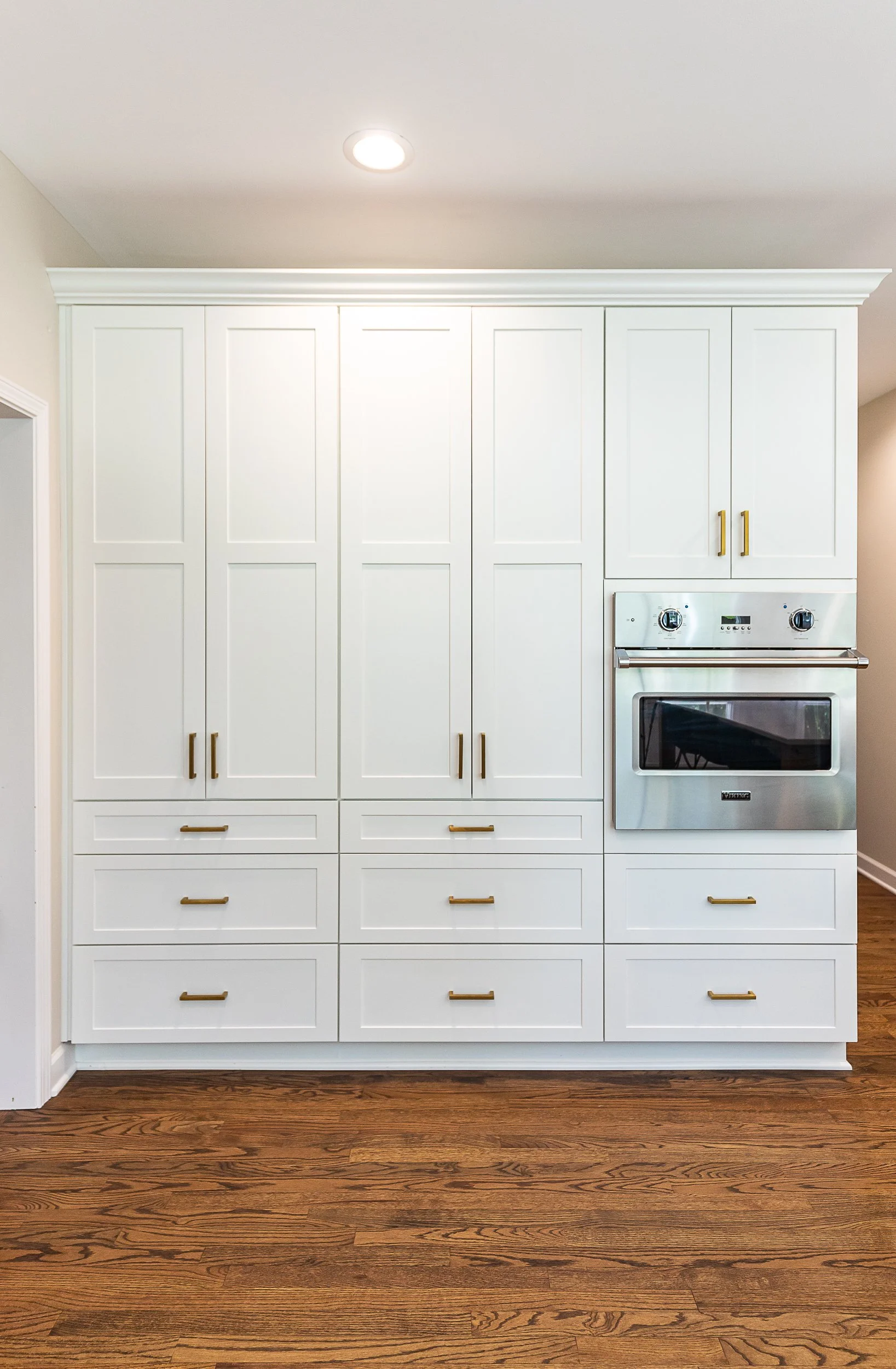 White kitchen wall with built-in cabinets and a stainless steel oven, wooden floor, ceiling light