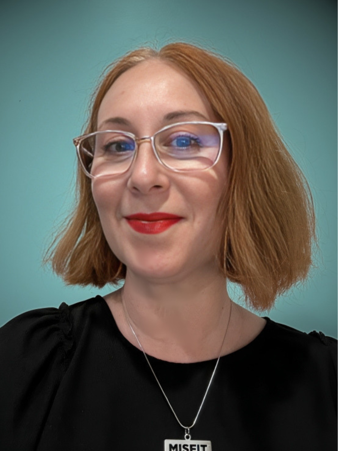 A woman with shoulder-length red hair wearing glasses, red lipstick, a black top, and a necklace with a pendant that reads 'MISFIT'.