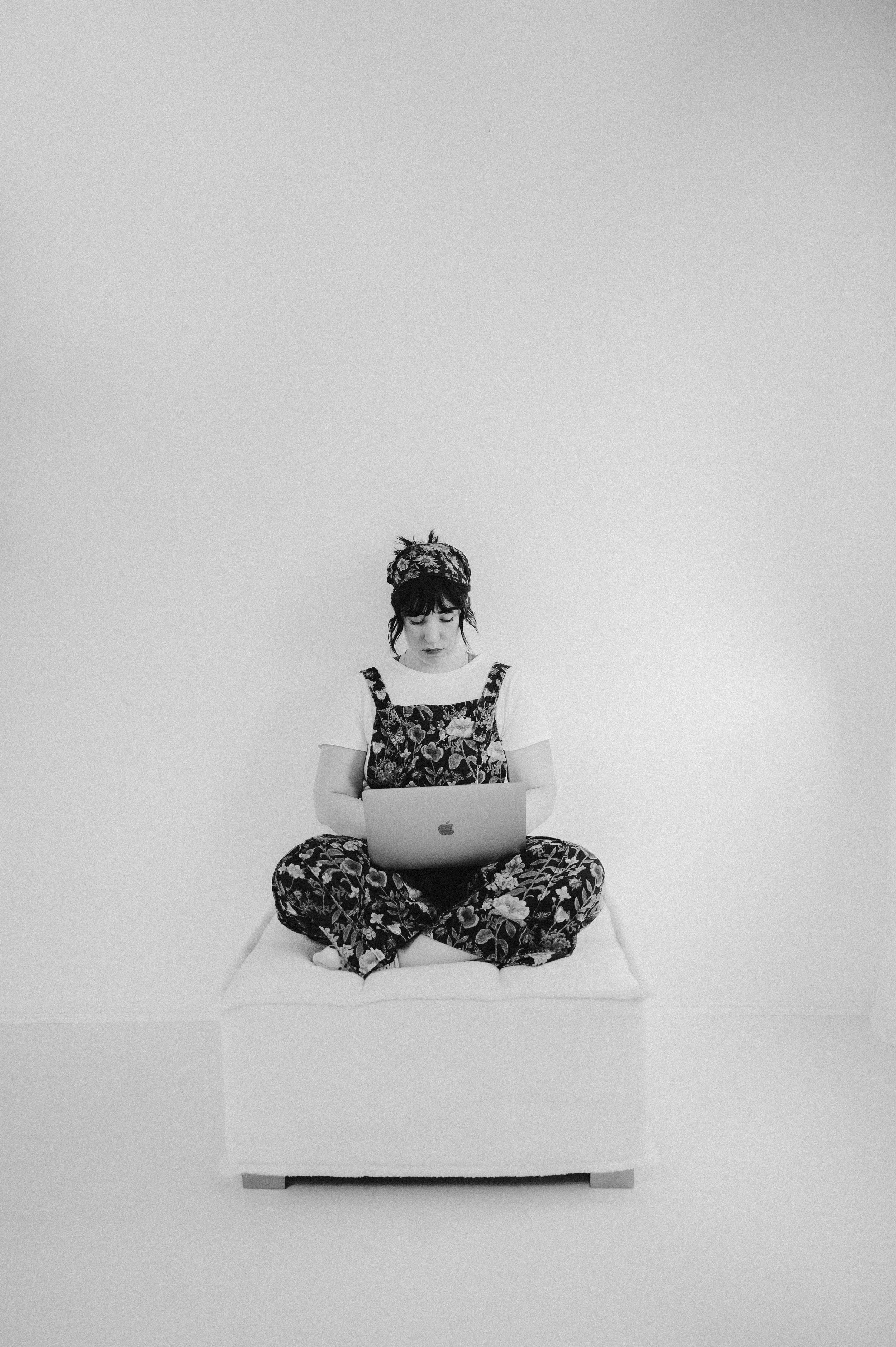 A young woman with dark hair, wearing a floral dress and a headscarf, sitting cross-legged on a padded platform in a minimalist white room, working on a laptop.